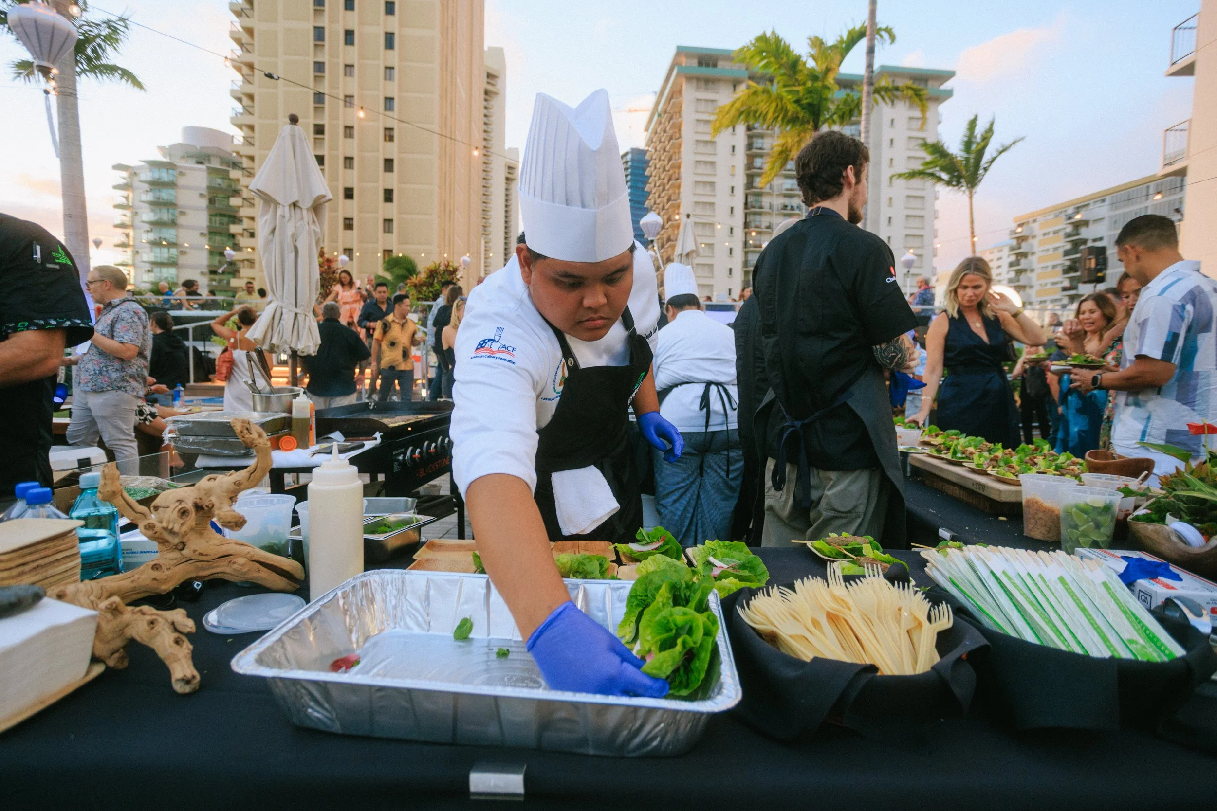 Chef preparing food at an outdoor event with people gathered around, tall buildings, and palm trees in the background.