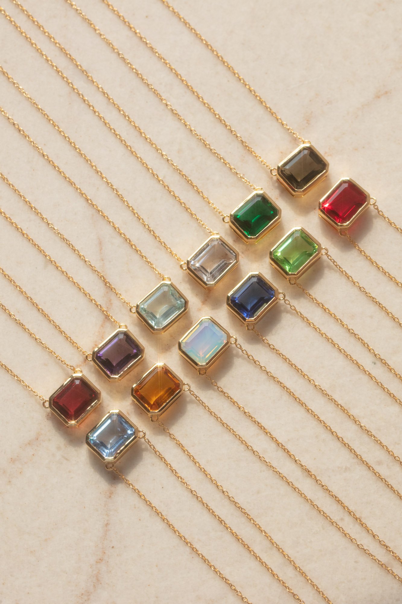 12 square shaped pendants sit in a line up row on yellow marble. They represent the 12 months of birthstones and are lined up in chronological order.