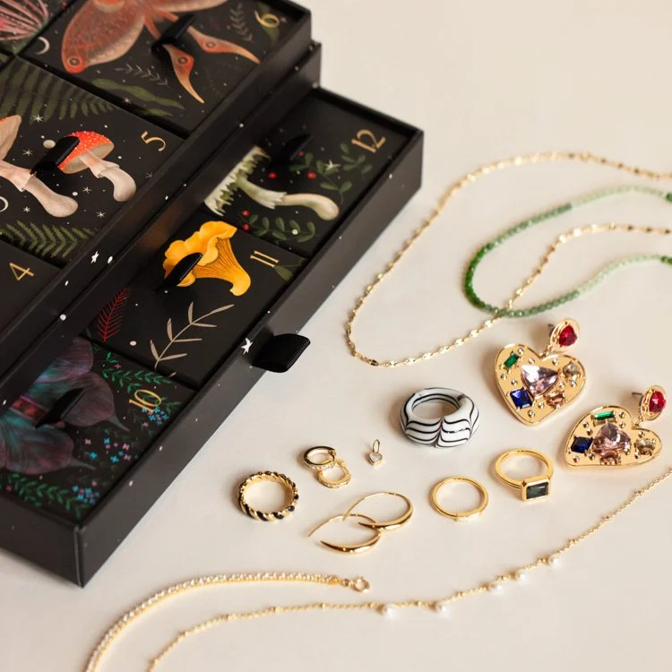 Jewelry from the Local Eclectic Advent Calendar lays on tan backdrop next to the box. They are yellow gold rings, necklaces, anklets and earrings.