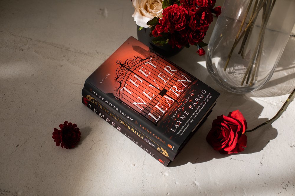 "They Never Learn", a book by Chicago author Layne Fargo sits on top of other books on a white cement floor. There are red roses on the floor and in a vase next to them.