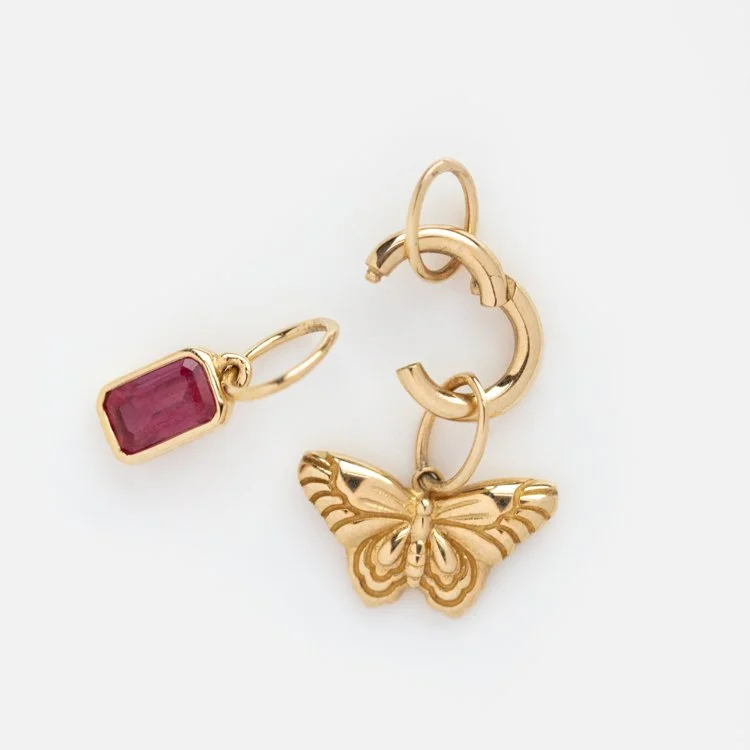 A ruby red charm with a yellow gold setting sitting next to a charm clip with a butterfly charm on it.