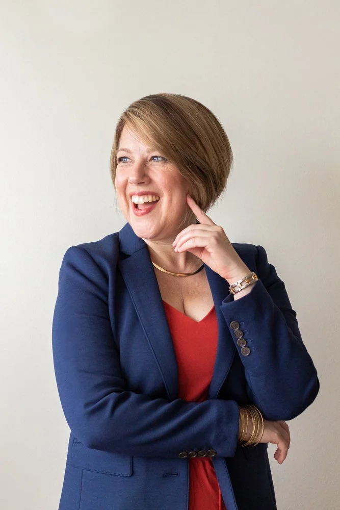 A woman in a blue suit jacket with short brown hair against a white wall