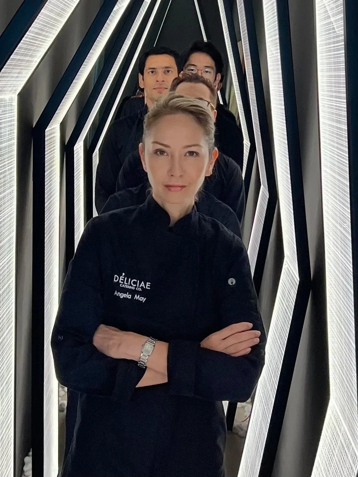 A group of people in black chefs uniforms standing in a line in a modern, illuminated corridor. The woman at the front has a watch on her left wrist and a name tag that reads 'Angela May' and 'DÉLICIAE Catering Co.'