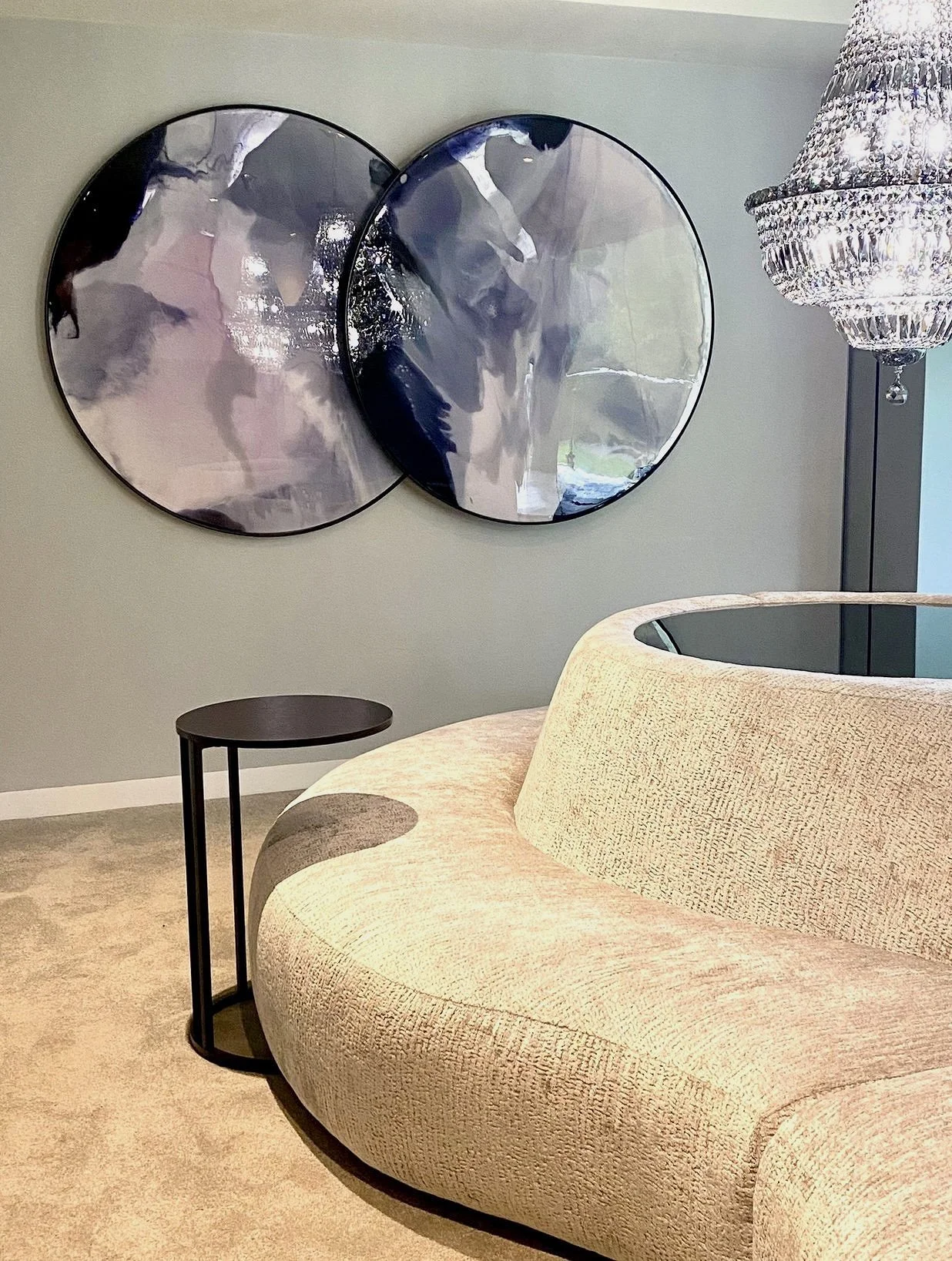 Modern living room corner with beige textured curved sofa, round black side table, abstract wall art with two circular metallic pieces, and a crystal chandelier hanging from the ceiling.