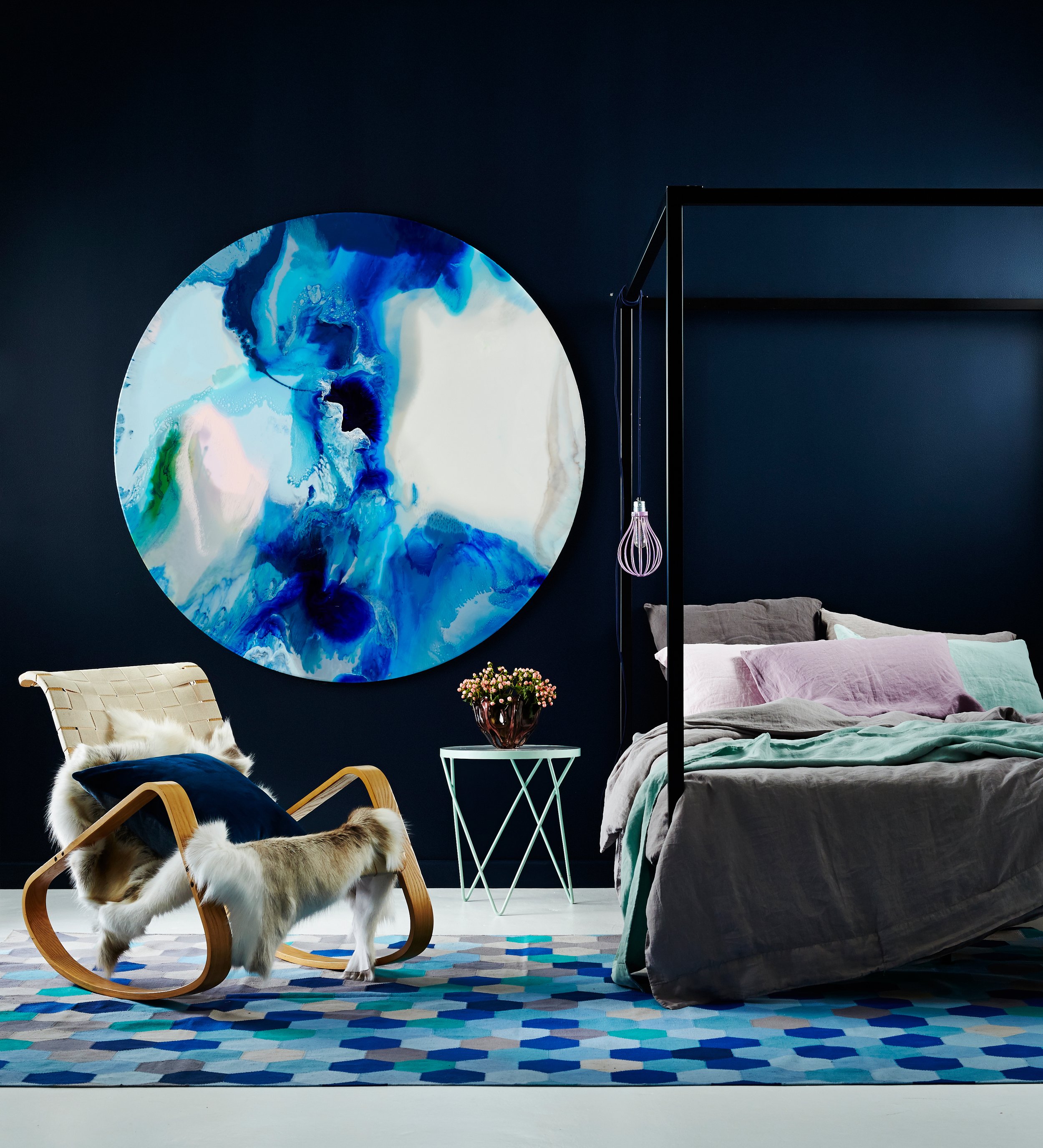 Modern bedroom with dark blue wall, large round abstract art with blue, white, and green swirls, bed with gray bedding and pastel pillows, black metal canopy frame, wooden rocking chair with fur throw and navy cushion, small white side table with a p