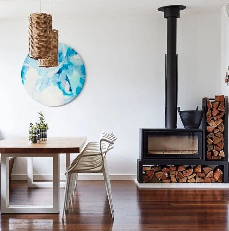 Modern dining room with wooden table, white chairs, and a black wood stove with stacked firewood, a blue and white abstract art piece on the wall, and hanging wicker lamps
