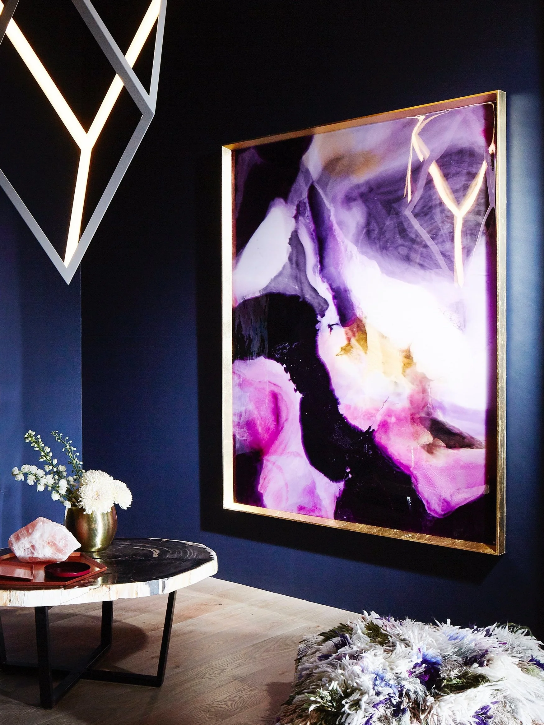 An abstract purple, black, and white painting on a dark wall, illuminated by a neon light in a geometric shape, with a small table holding flowers, a large crystal, and a decorative tray, and a fluffy white and purple rug on the floor.