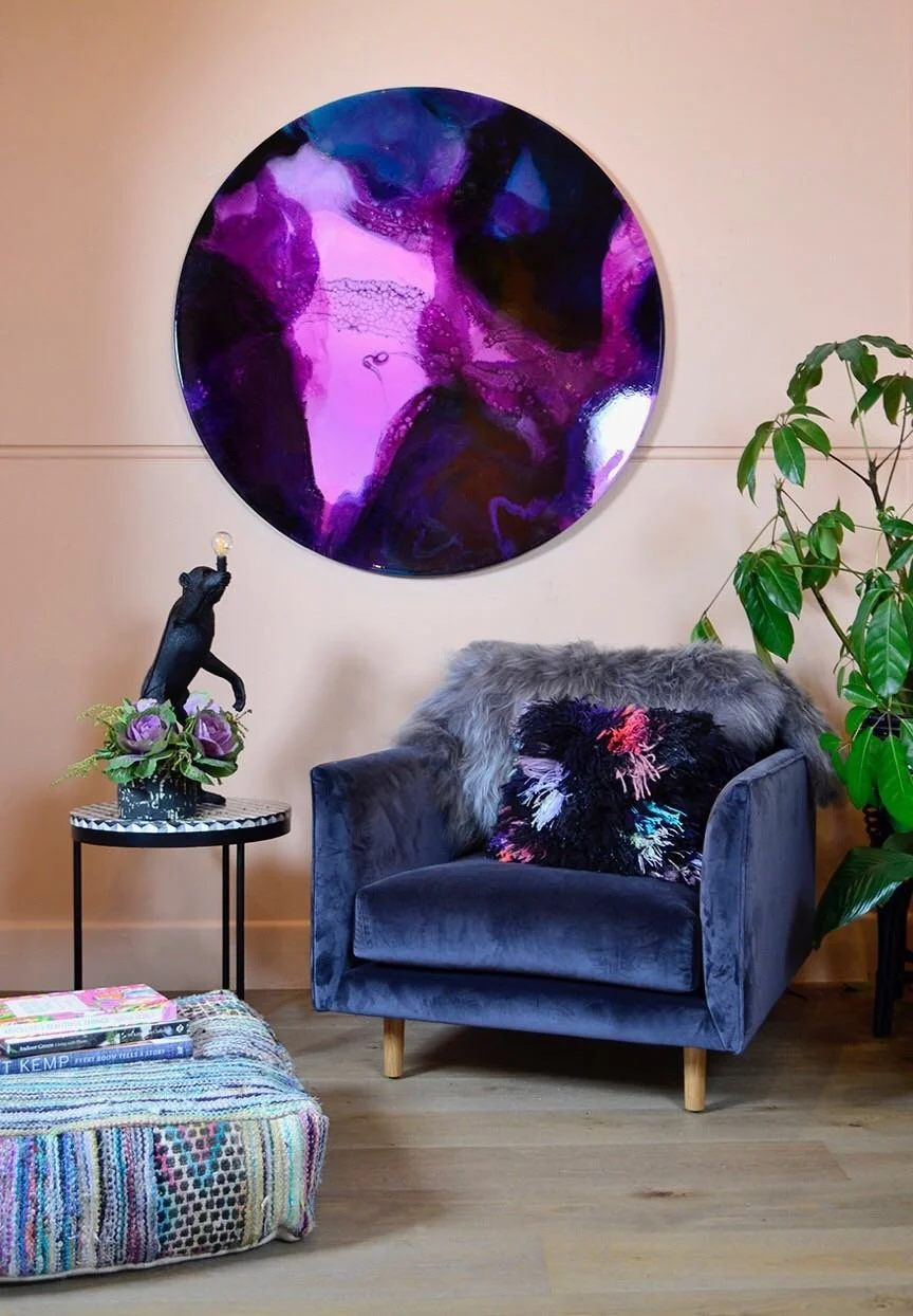 A cozy living room corner featuring a blue velvet armchair with a colorful abstract pillow and faux fur throw, a small side table with a black bear sculpture holding a light bulb, a pink and purple flower arrangement, a large round purple and black a