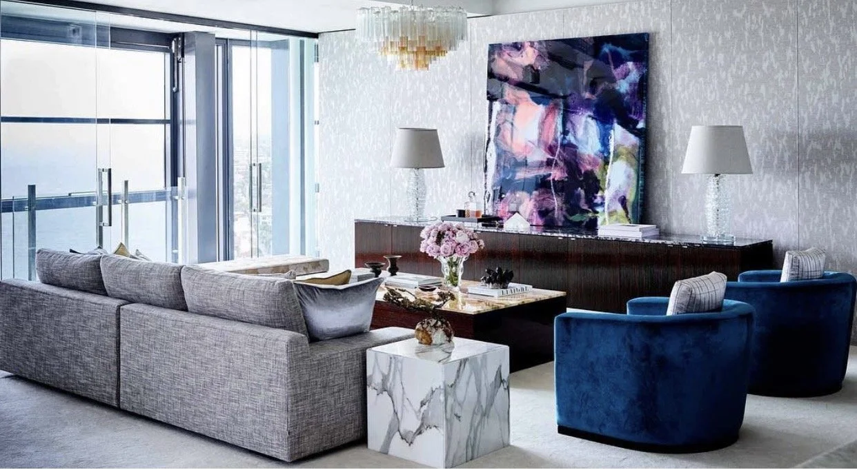 Modern living room with gray sofa, blue armchairs, marble side table, floral centerpiece, abstract wall art, and large windows.