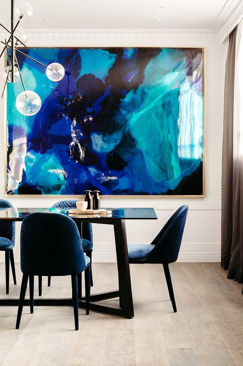 Modern dining room with a square black table, four navy velvet chairs, abstract blue and teal painting on white wall, and a chandelier with glass globes.