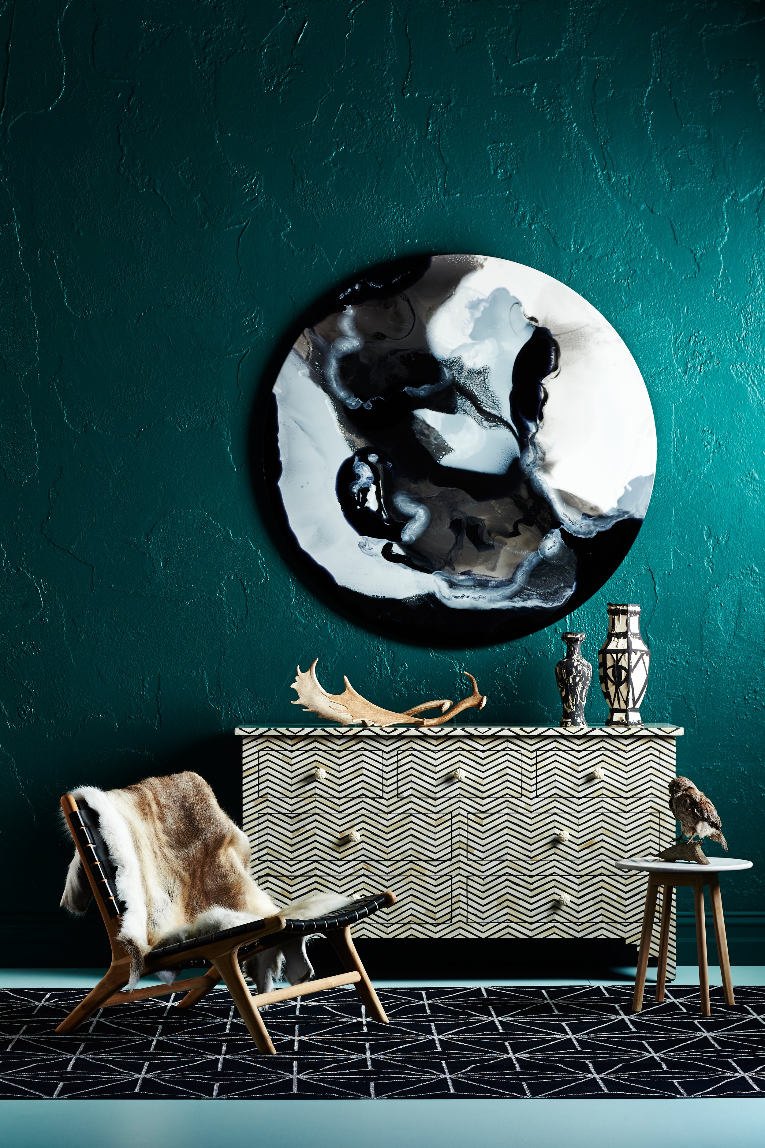 Modern interior with a teal textured wall, a black and white abstract wall art, a patterned black and white sideboard, a wooden chair with a fur throw, and decorative vases and sculptures.