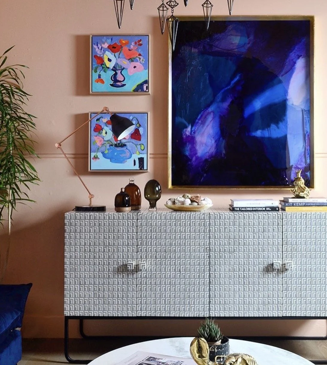 Decorative living room wall with abstract artwork, colorful paintings of flowers, a modern desk lamp, vases, stacks of books, small sculptures, and a patterned sideboard, with a small plant and art pieces on a white table in foreground.