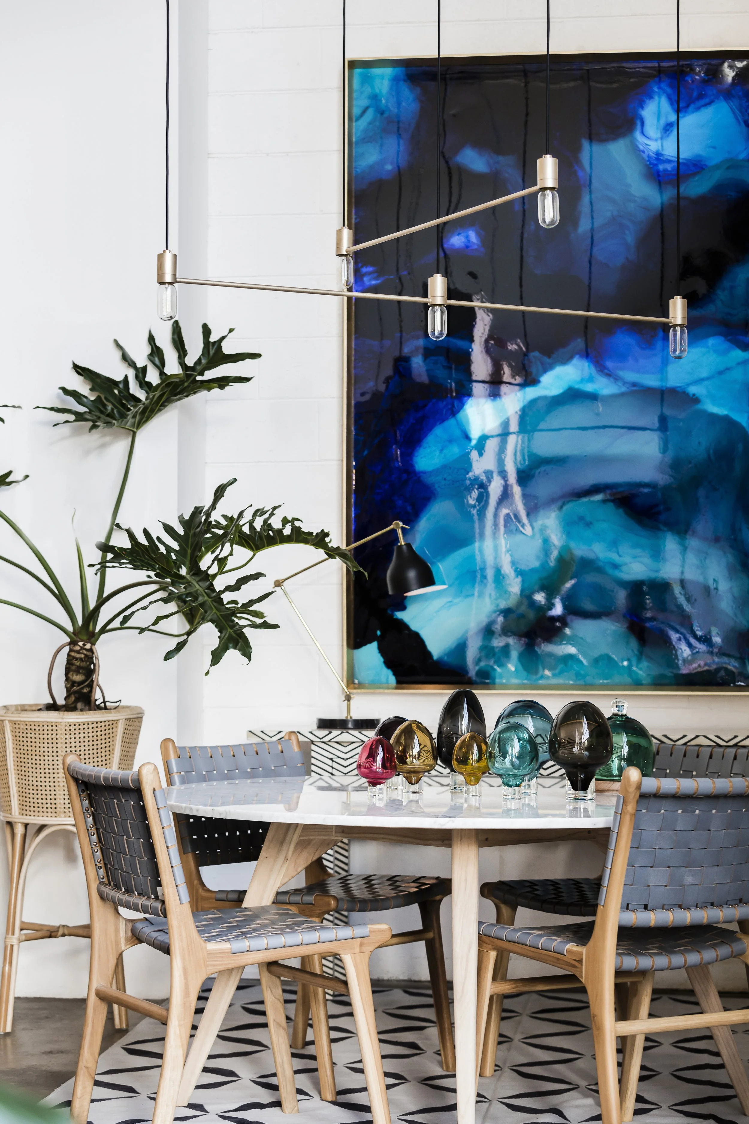 A dining area featuring a round table with colorful glass vases, six woven chairs with gray seats, an abstract blue artwork on the wall, a black desk lamp, and a large green plant in a wicker pot.
