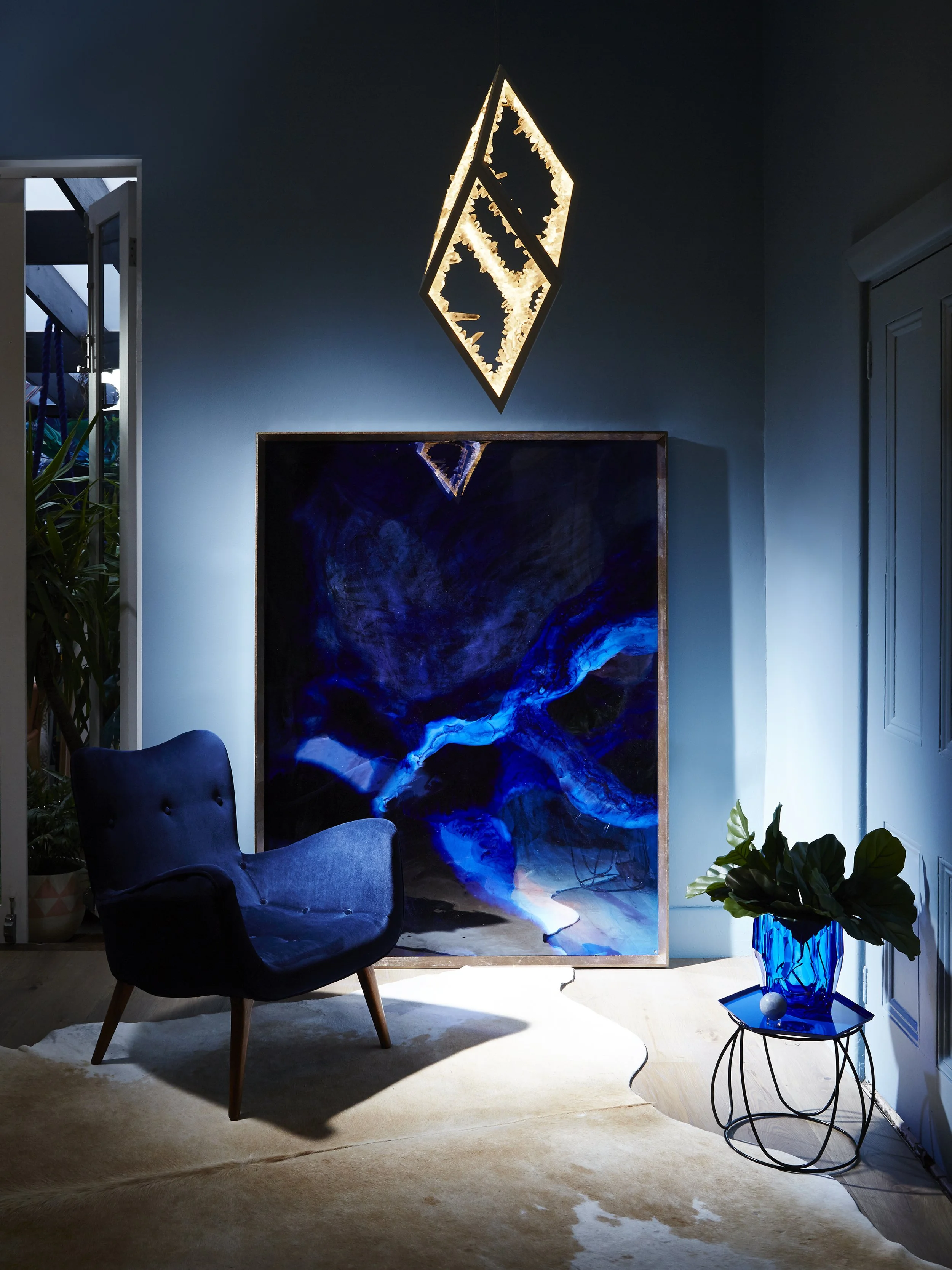 A cozy interior with a blue armchair, abstract blue and black artwork, a small round black table with a blue vase containing green leaves, a white ball, and a decorative wall light shaped like a diamond hanging from the ceiling