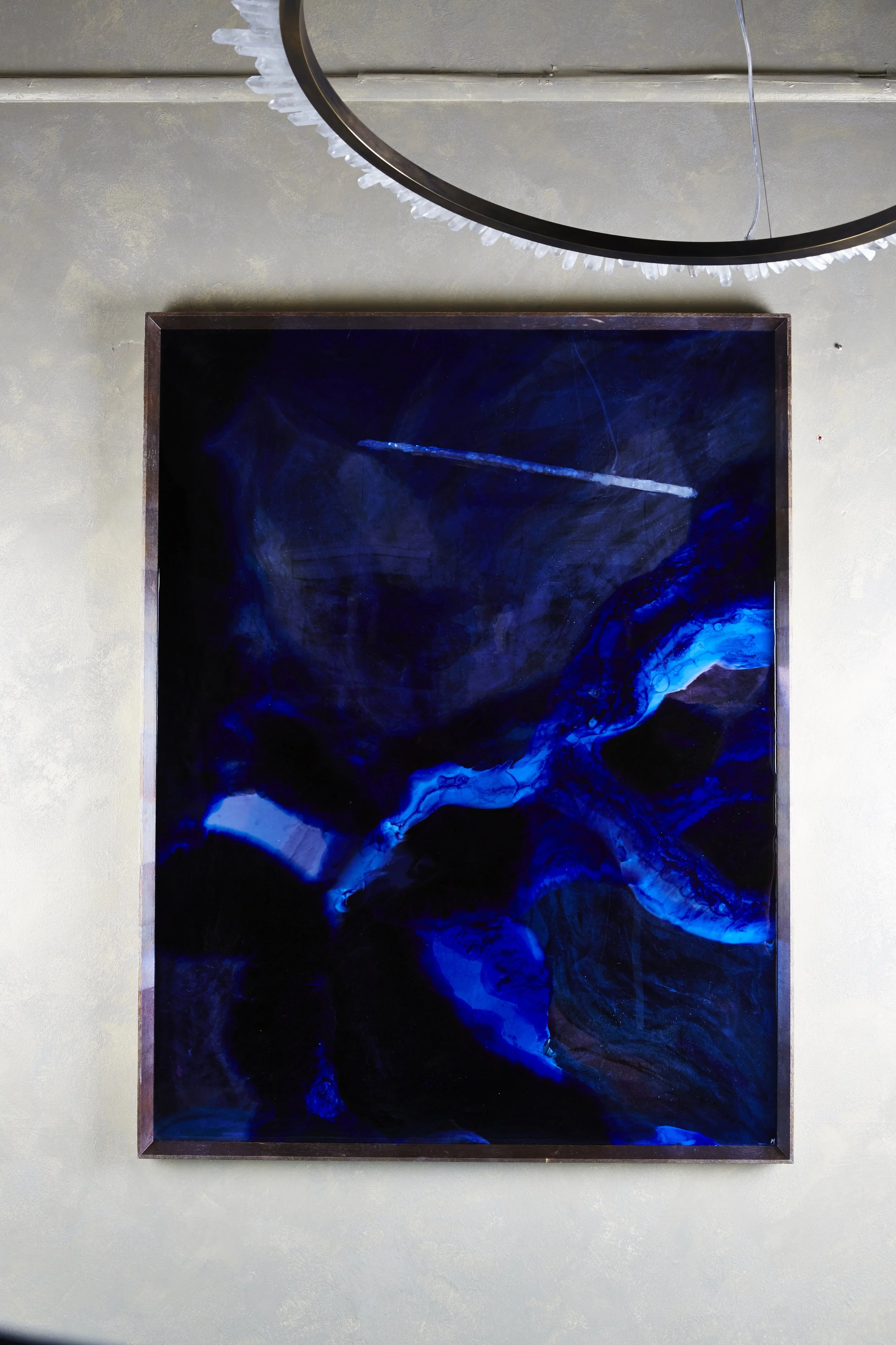 An abstract painting with swirling shades of dark blue, light blue, and black, framed in wood, hanging on a beige wall.