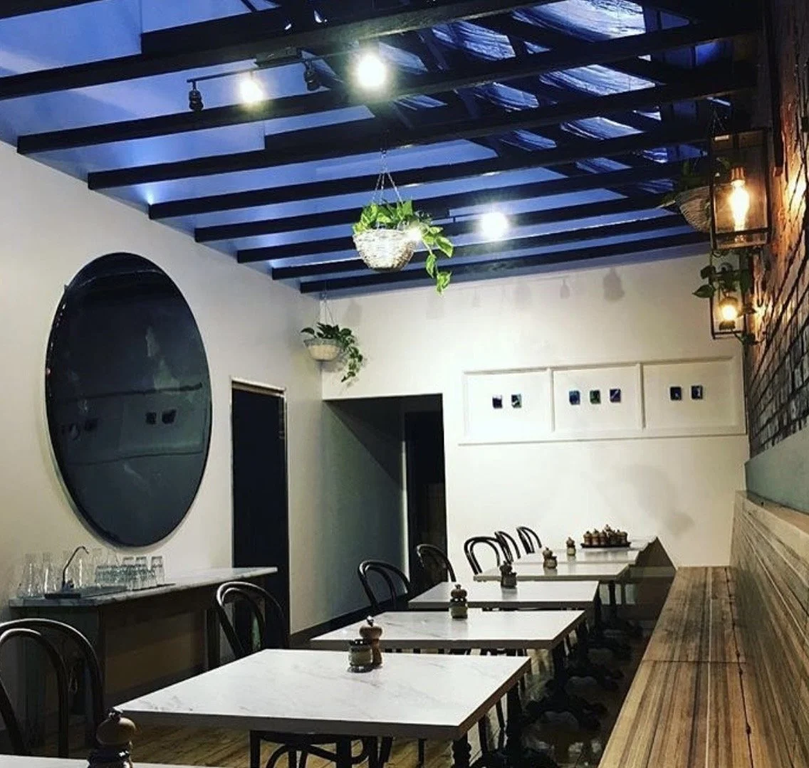 Interior of a restaurant or cafe with white marble tables, black chairs, wooden accents, hanging potted plants, wall-mounted lantern-style lights, and a large circular mirror on the wall.
