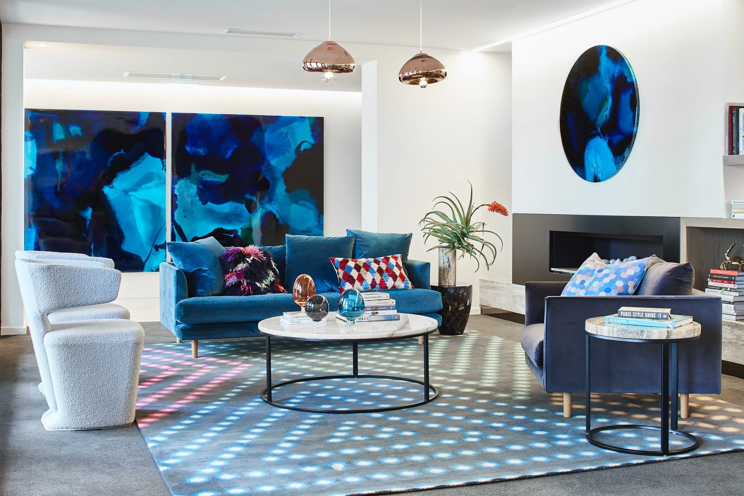 Modern living room with blue sofas, abstract blue artwork on the wall, white and black side tables, colorful decorative pillows, potted plant, and patterned rug.
