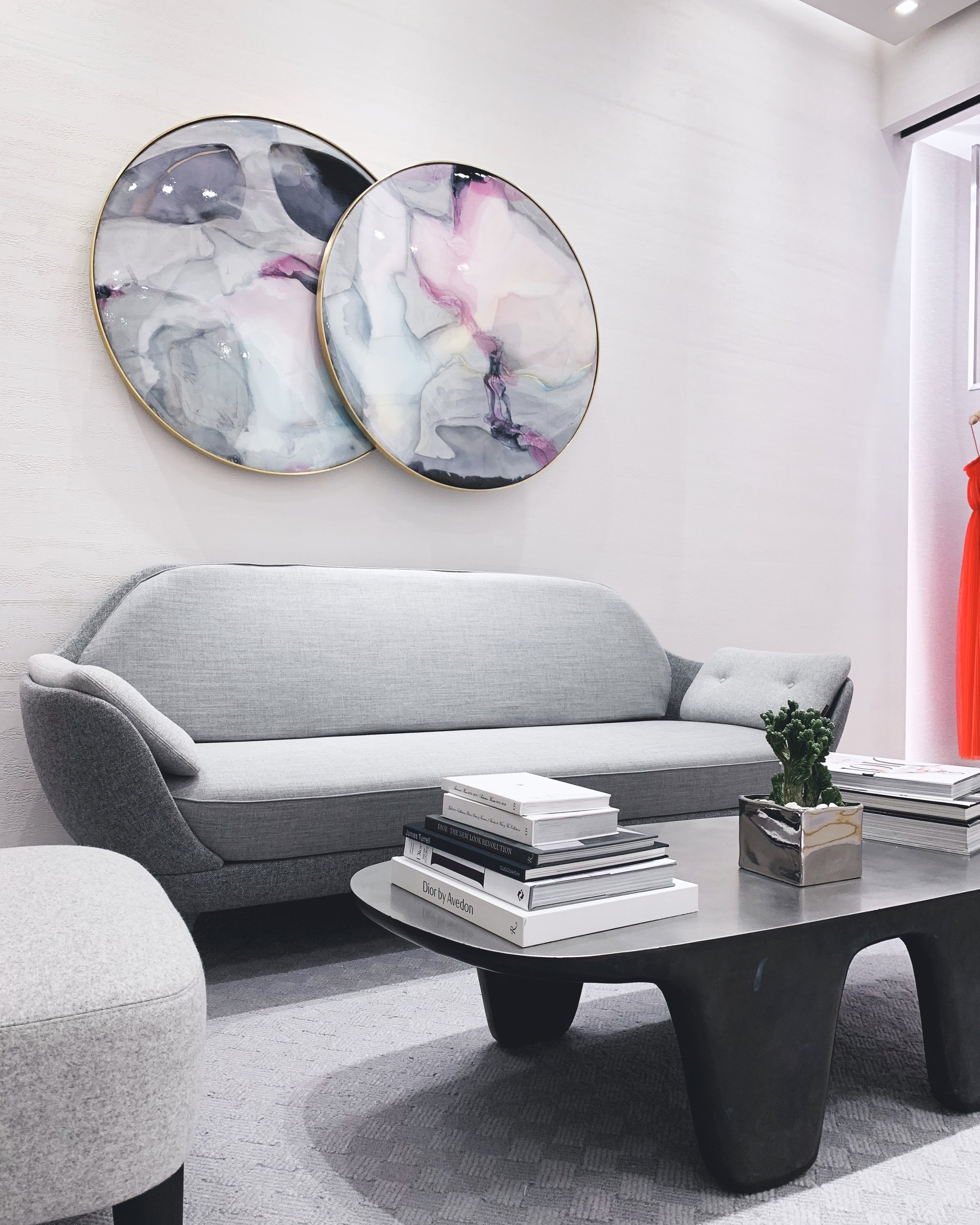 Dior vip room with a light gray sofa, abstract circular wall art above, a black coffee table with stacked books and a small potted cactus, and a red curtain partially visible on the right side.