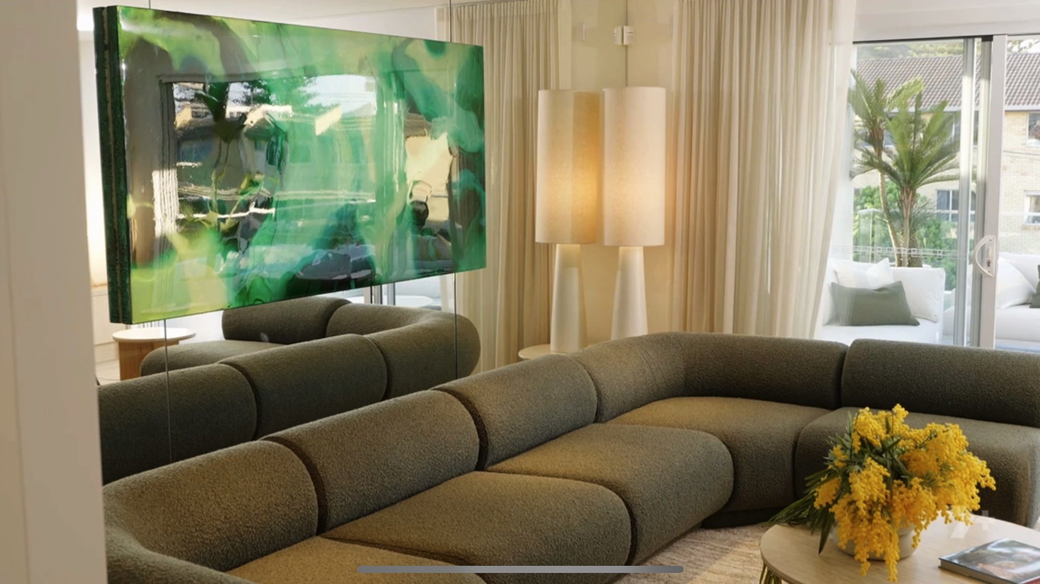 A modern living room with a large green artwork on the wall, a gray sectional sofa, a bouquet of yellow flowers on a coffee table, a tall floor lamp, and sliding glass doors leading to an outdoor patio with white outdoor furniture and a palm tree.