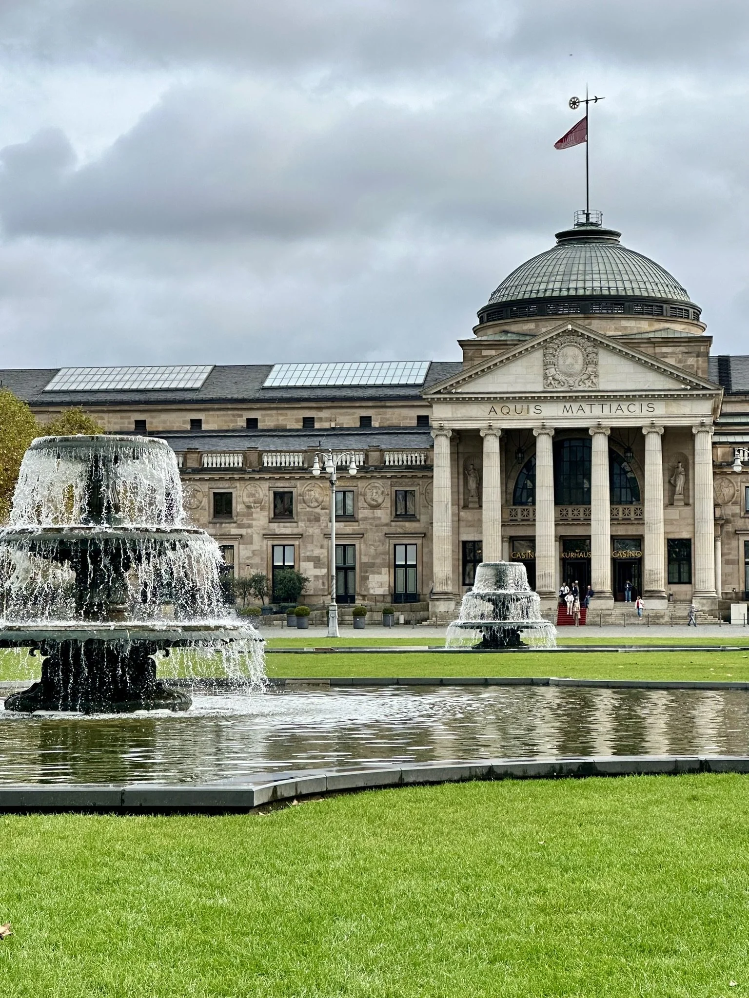 View of the Kurhaus, casion, in Wiesbaden, a historic building with classical columns and statues on the facade, a dome on top, and a flag flying from the roof. In front, a large fountain with cascading water.