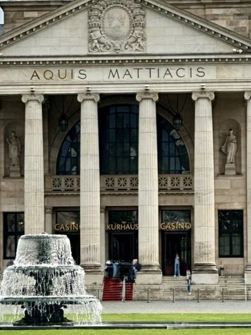 Front of a classical-style casino building in Wiesbaden, with tall columns, a fountain in front, and the phrase 'AQUIS MATTIAICS' inscribed above the entrance.