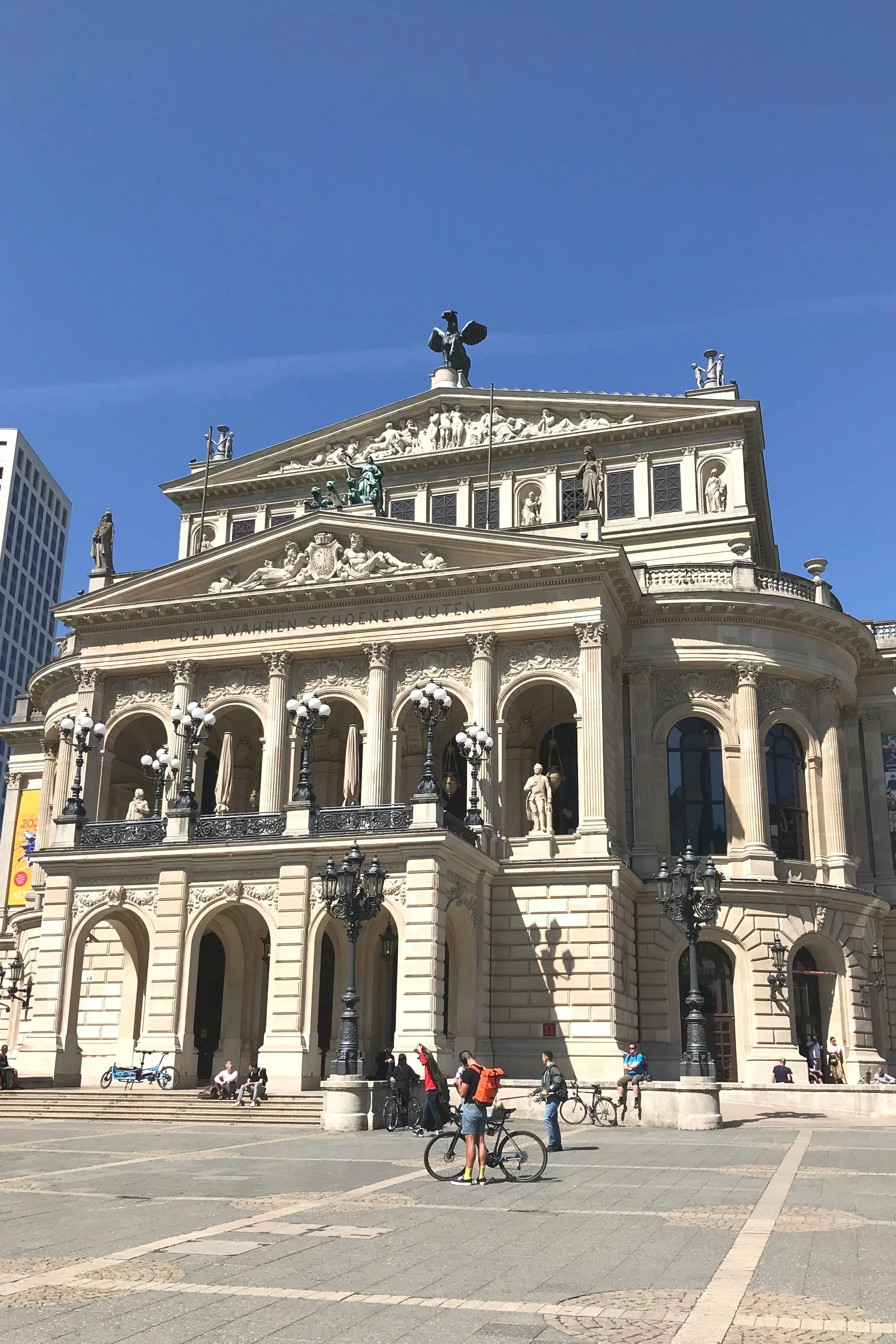 A grand, historic Old Opera House in Frankfurt with classical architecture, multiple statues, ornate columns, and decorative details, situated in an open plaza with people, bicycles, and modern buildings nearby.
