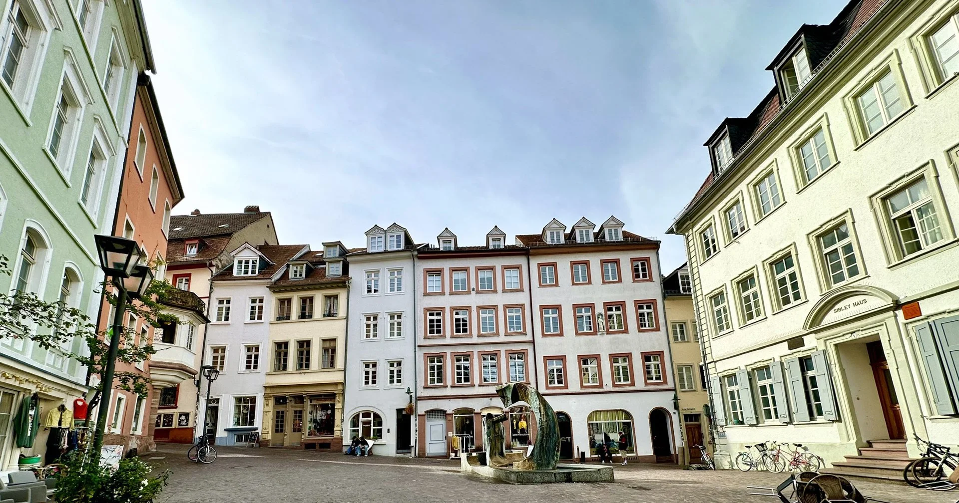Heidelberg Heumarkt, colorful, baroque, European buildings surround a small cobblestone square with a modern sculpture fountain, bicycles, and a few people sitting and walking.
