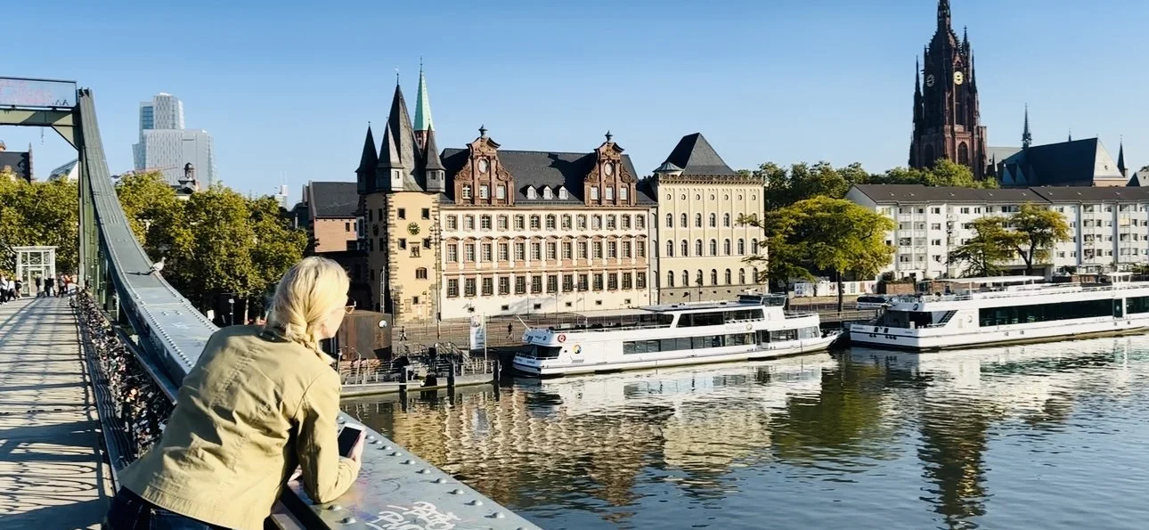 Anne Noble leaning on the Eisernersteg, Frankfurt, a bridge looking at boats on the River Main with historic museum building and cathedral dome in the background.