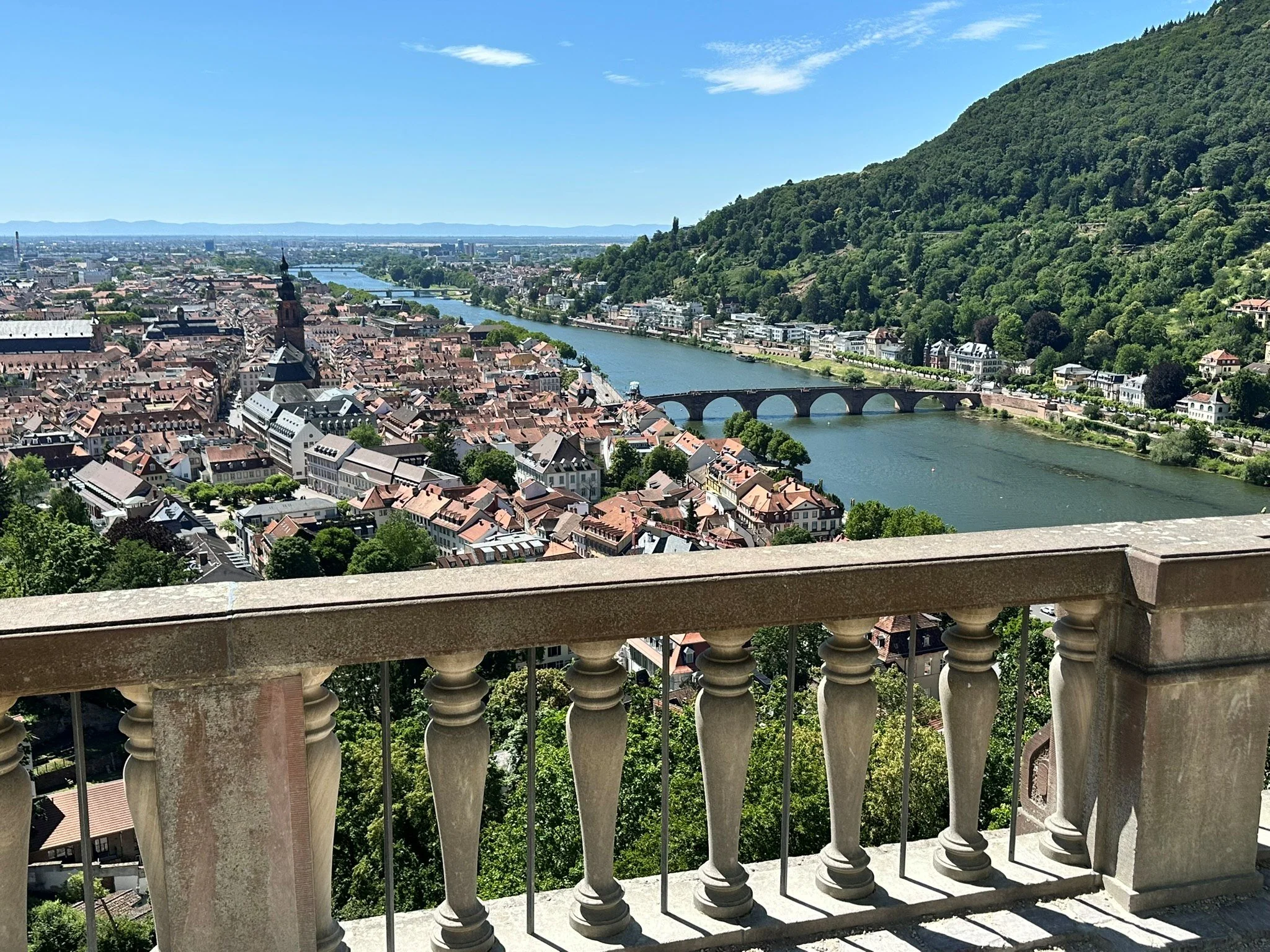 Aerial view of Heidelberg with historic buildings, a river with an arched stone bridge, and a green hillside in the background, viewed from Heidelberg castle balcony with stone balustrades.