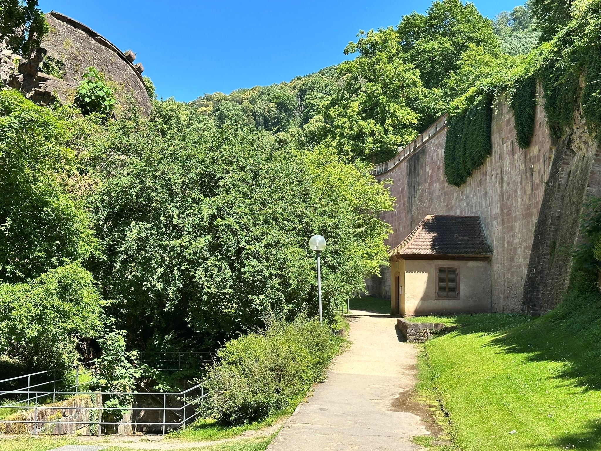 A small fountain building with shutters on the windows situated next to a tall stone wall in Heidelberg castle moat. A pathway runs past the building, with greenery and trees on either side, under a bright blue sky.