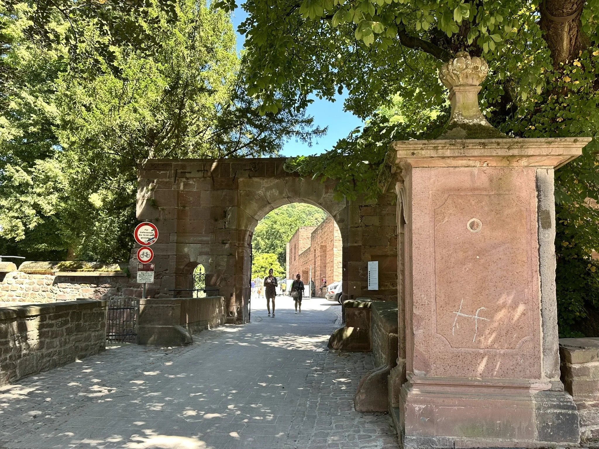 Historic stone archway entrance to Heidelberg castle flanked by a pink sandstone pillar on the right with decorative top and a vintage plaque. Behind the arch, a stone wall and cobblestone pathway, with lush green trees lining the area.