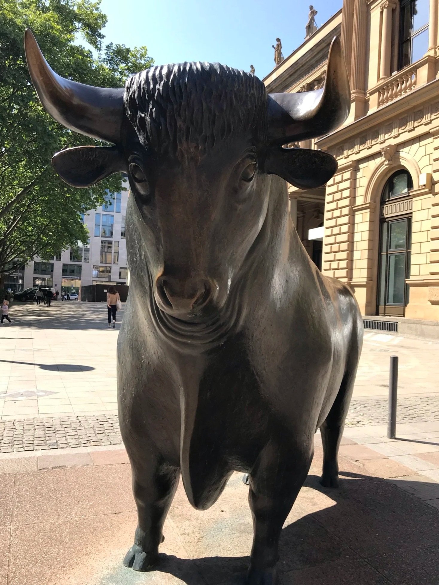 A large bronze statue of a bull standing outside the Frankfurt Stock Exchange with trees and buildings around.