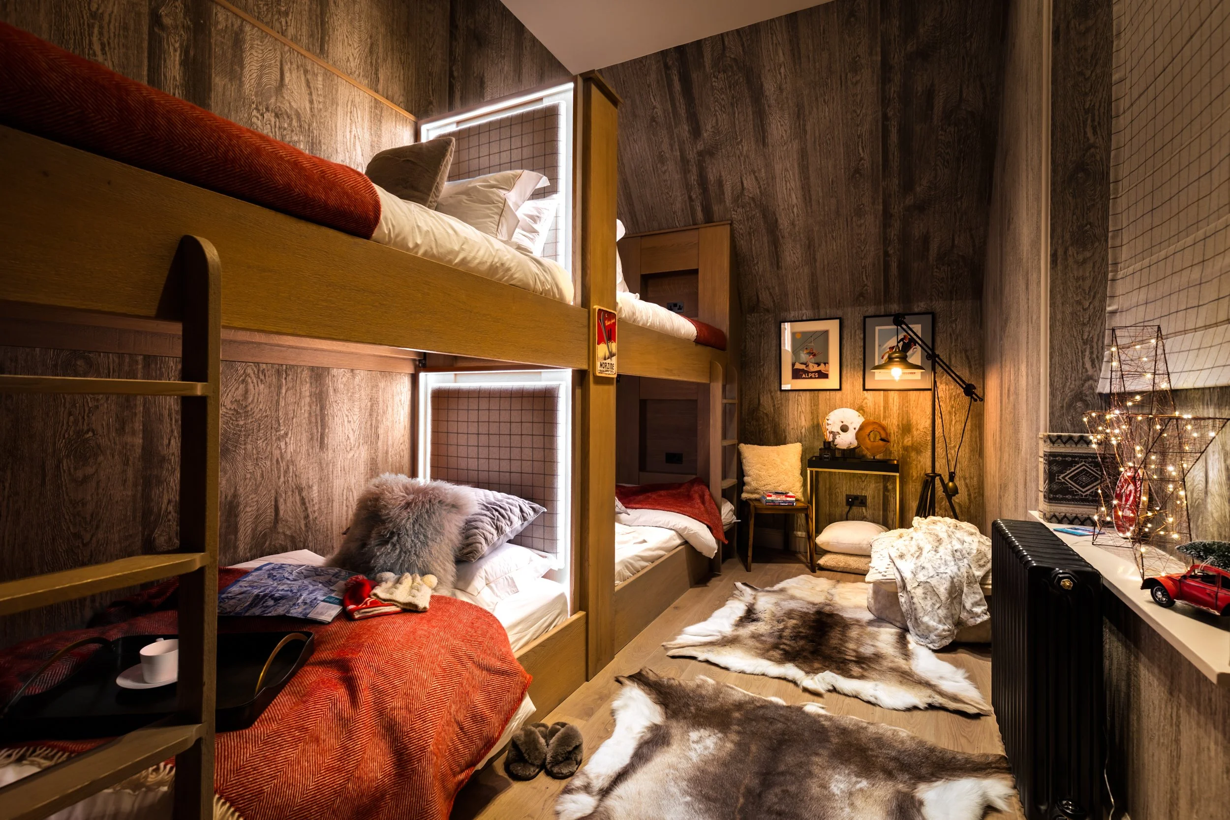 A cozy bedroom featuring a double bunk bed with wooden frames, surrounded by wooden-paneled walls and additional furnishings like chairs, rugs, and decorative items, illuminated with warm lighting.