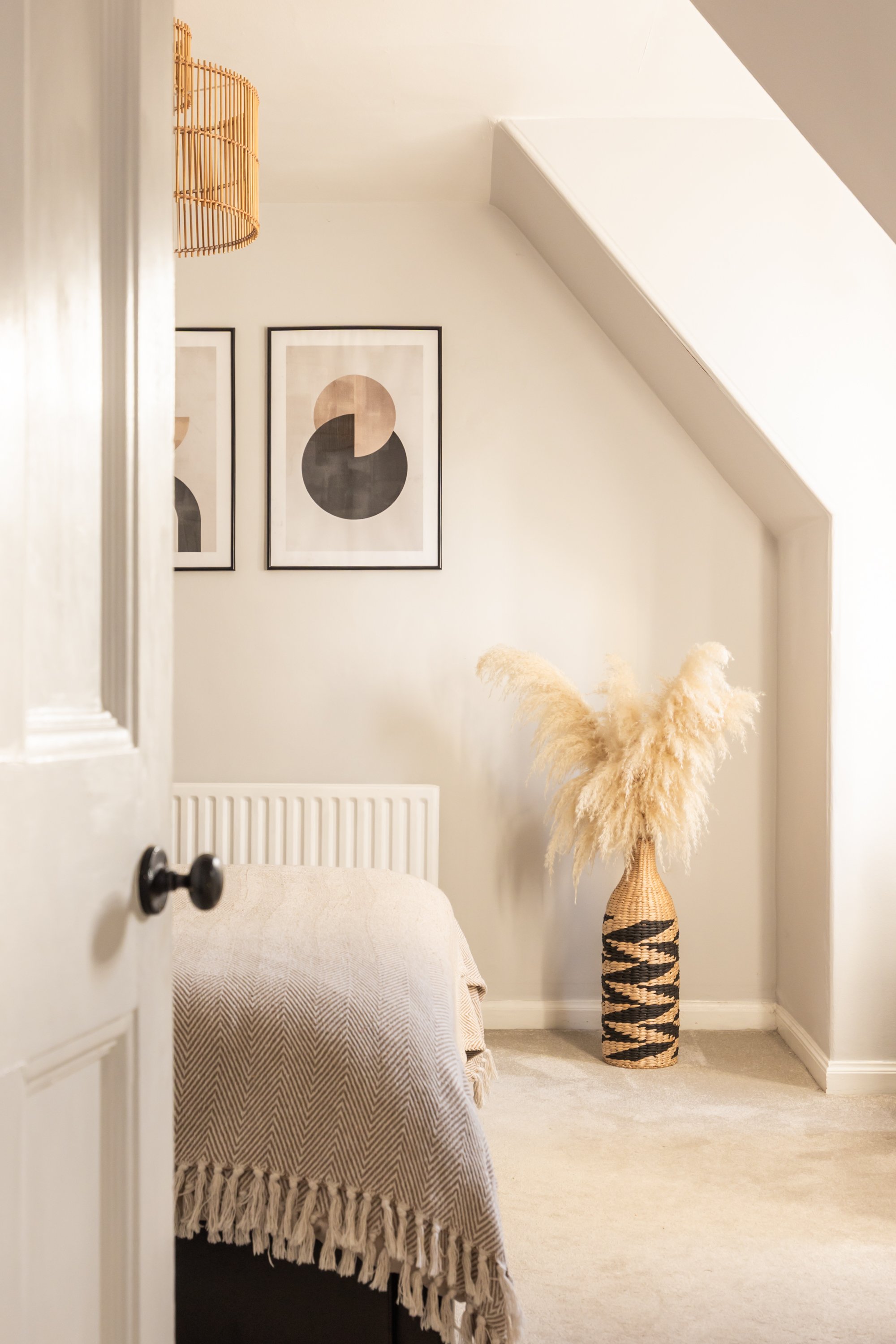 A cozy bedroom with a bed covered in a textured beige throw blanket, a tall woven vase with pampas grass next to a sloped wall, and minimalist abstract art on the wall.