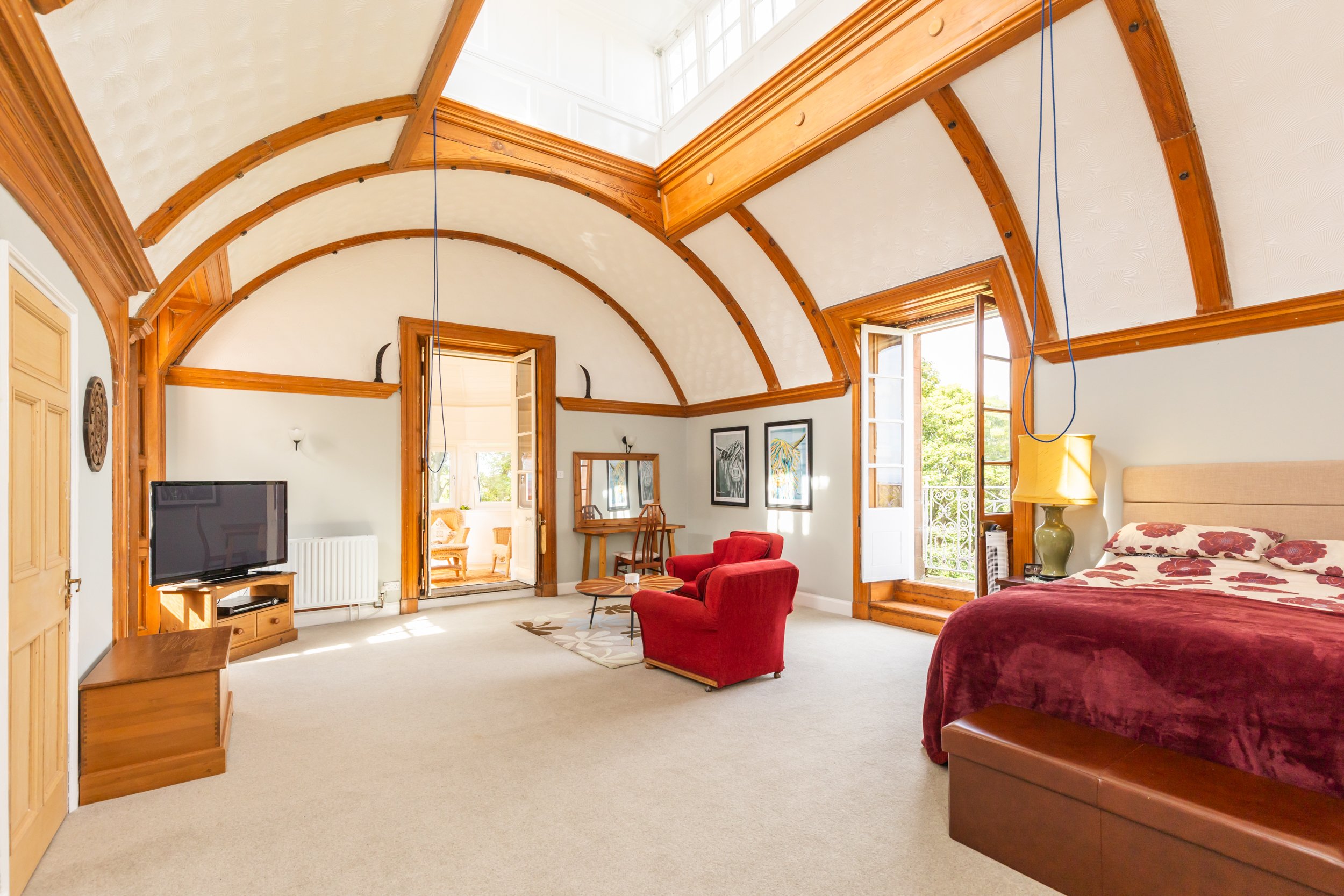 Room with vaulted ceiling and wooden accents, featuring a bed with red bedding, a red armchair, a TV, and balcony doors opening to greenery.