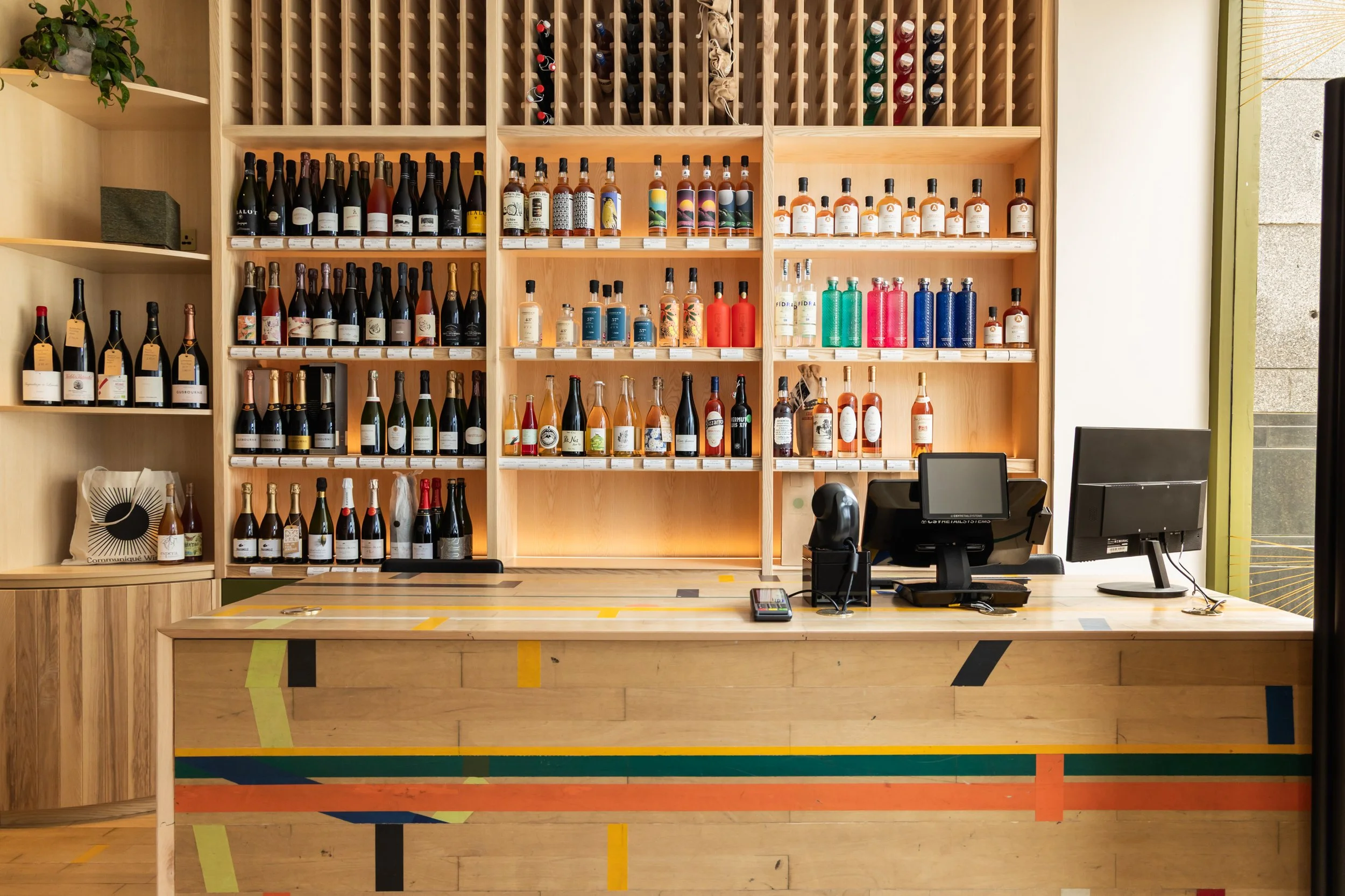 A wine store checkout counter with a colorful geometric design, behind which is a wooden shelving unit filled with bottles of wine and colorful liquor bottles, along with wine racks on top.
