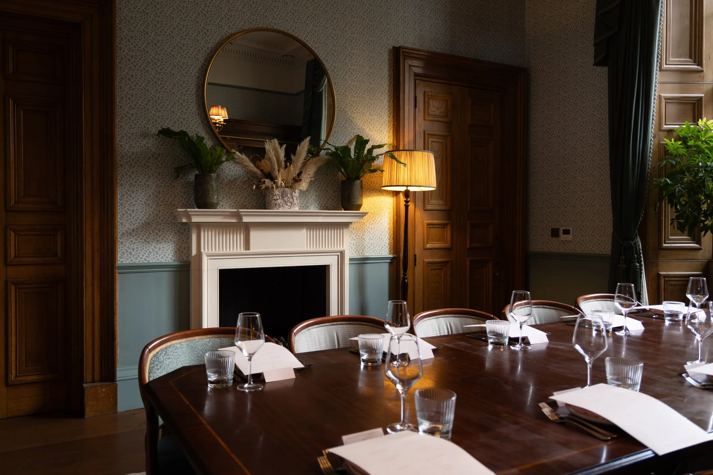 Formal dining room with a large wooden table set with glassware and napkins, a white fireplace with decorative vases and plants on mantel, a round mirror above, wooden paneling, and a tall window with dark curtains, illuminated by a floor lamp.