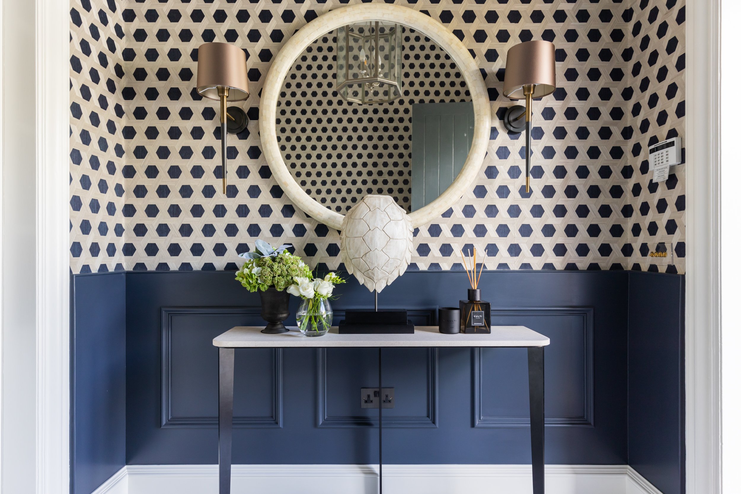 A decorative entryway with blue wainscoting and a patterned wallpaper, a large round mirror, two wall-mounted lamps, a white side table with vases of flowers, a sculpture, and a reed diffuser.