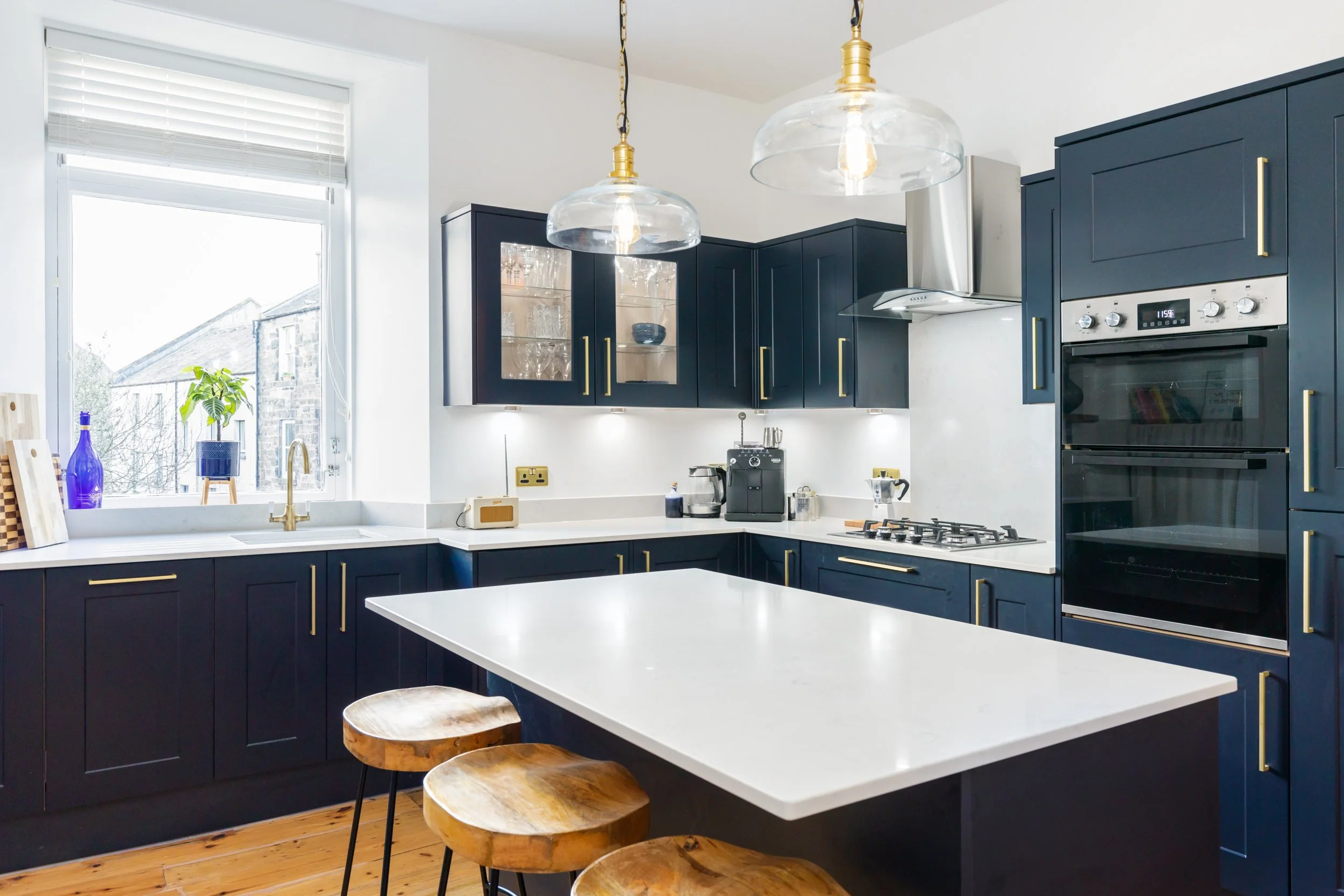 Modern kitchen with navy blue cabinets, white countertops, and a white island with wooden stools. Features include a large window, brass hardware, pendant lights, and built-in appliances.