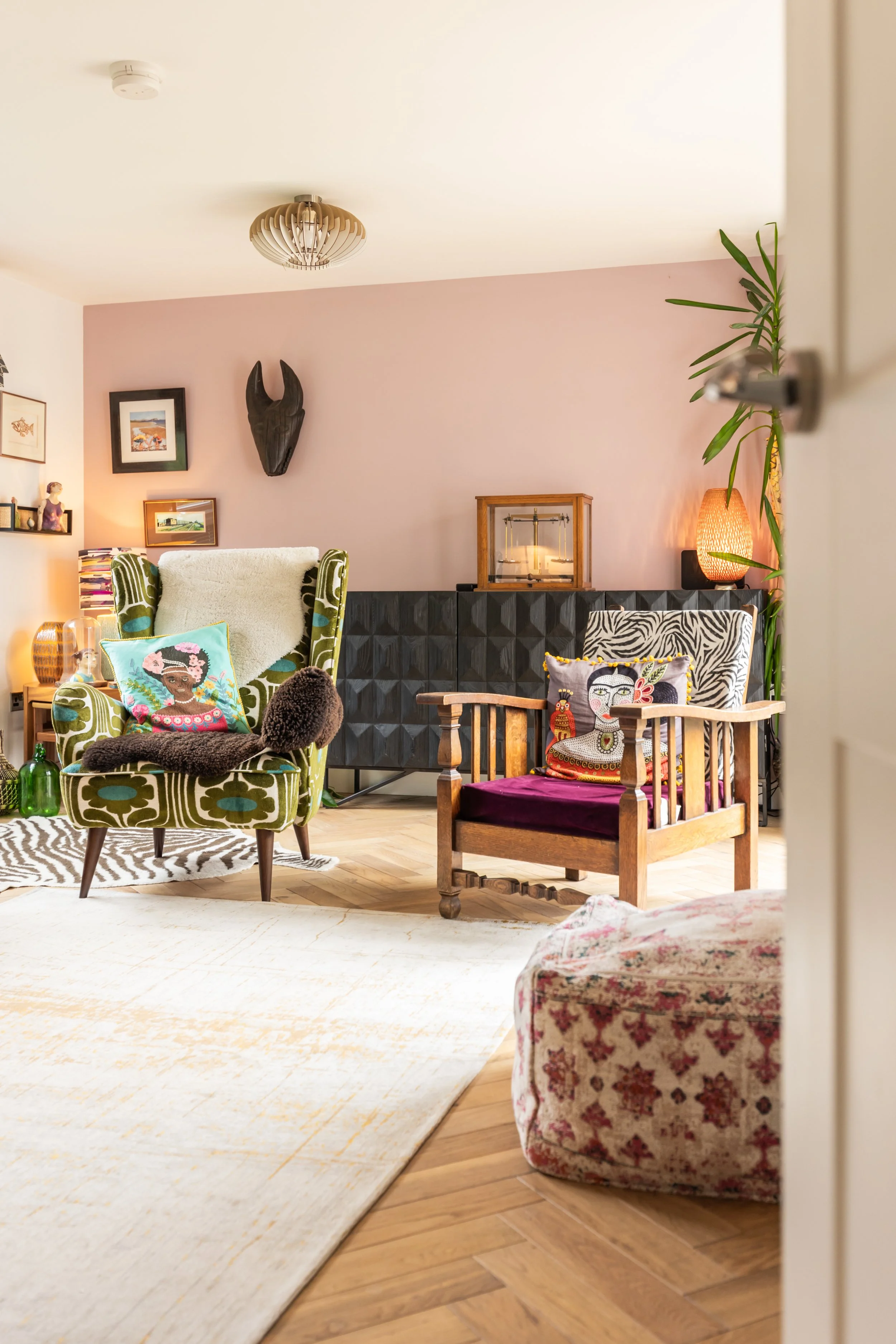 A cozy living room with vintage and eclectic furniture, including patterned armchairs, a wooden chair, colorful pillows, a textured lamp, potted plants, and framed artwork on the wall.