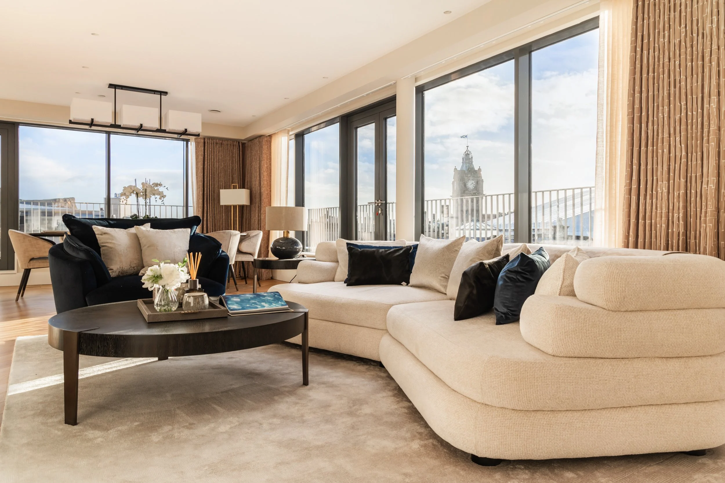 Modern living room with large windows, a beige sectional sofa with black and beige pillows, a dark oval coffee table with decorative items, and cityscape views including a clock tower.