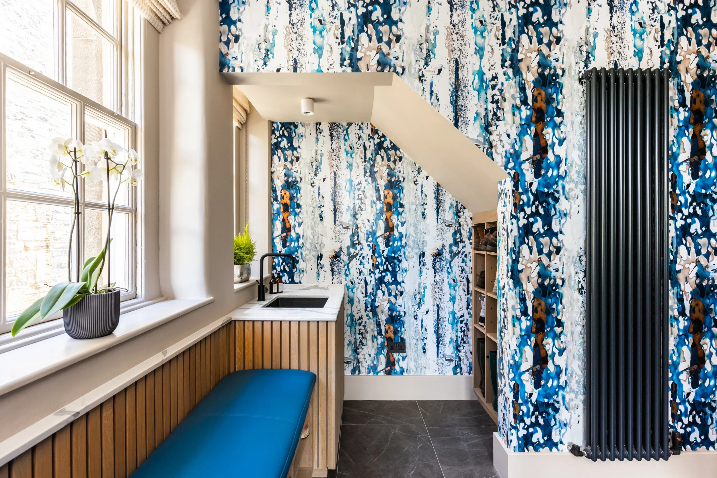 Interior of a modern kitchen corner with a window, white curtains, a black sink, a small countertop, an orchid plant, a green potted plant, a blue bench, and a vibrant blue abstract patterned wall with a black radiator.