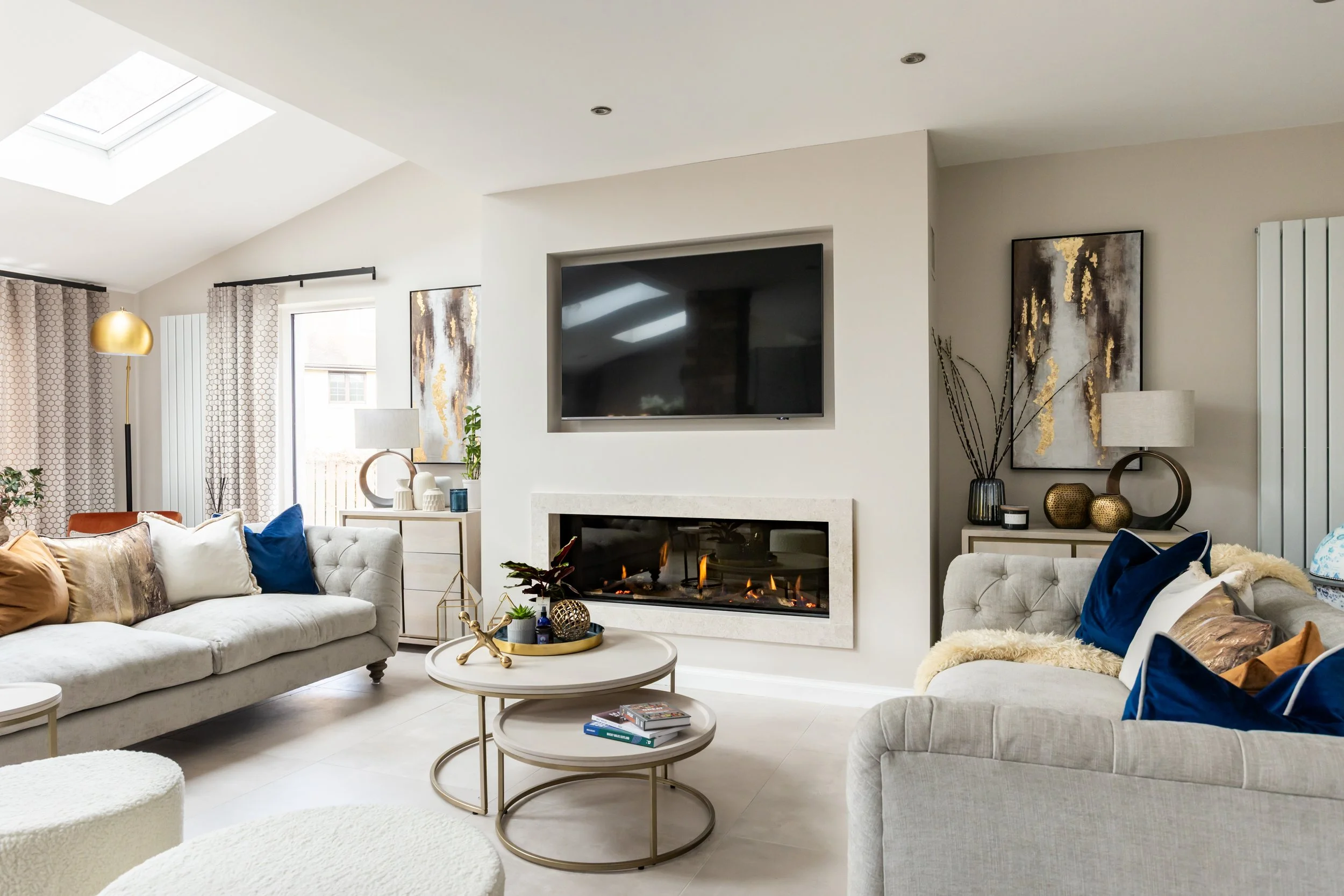 Modern living room with white walls, beige sofas with colorful pillows, a fireplace under a mounted flat-screen TV, a skylight, windows with curtains, and various decorative items including plants, lamps, and artwork.