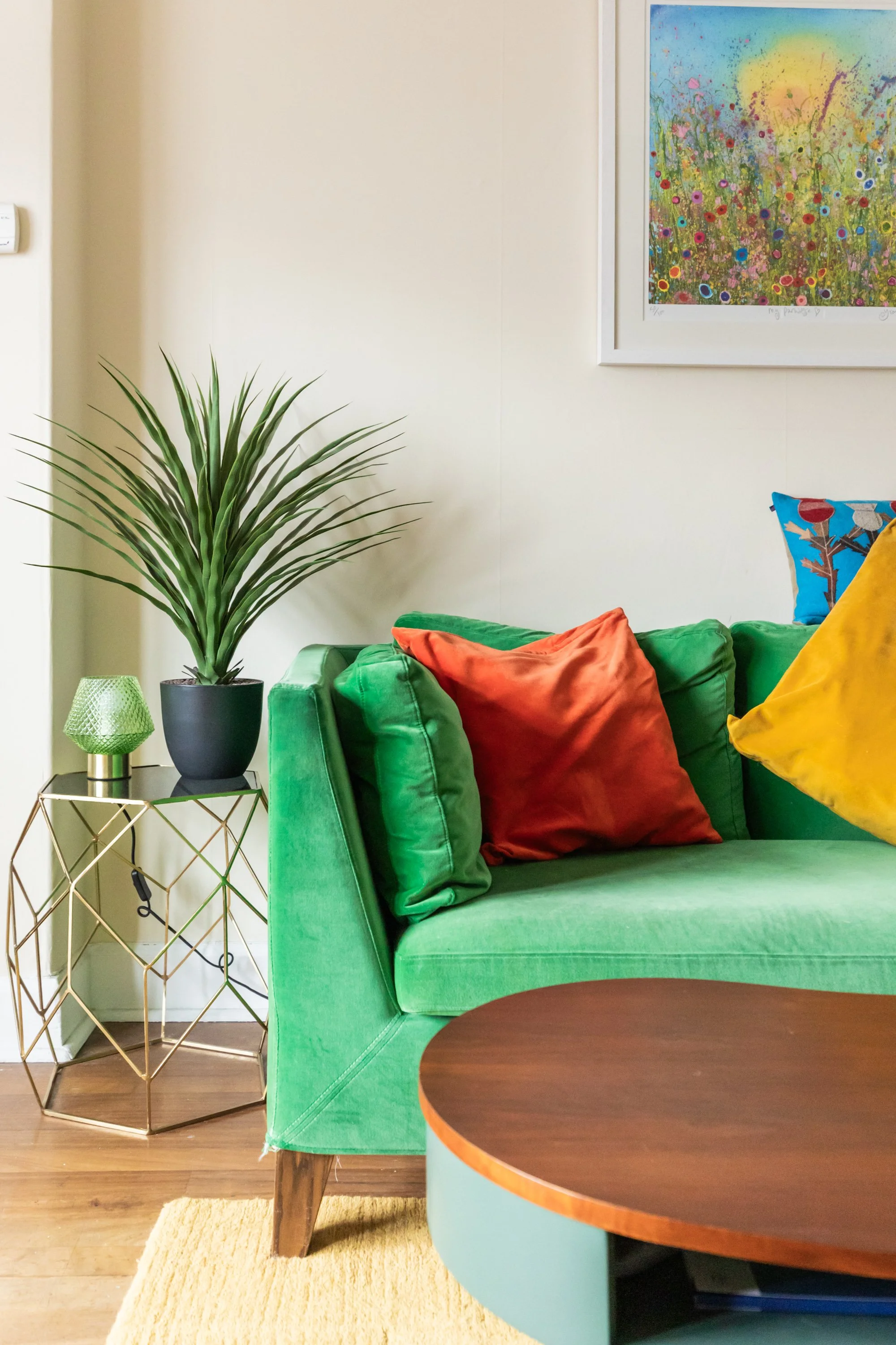 Colorful living room with a green sofa, orange, yellow, and blue pillows, a side table with a potted plant and a green lamp, wooden flooring, and a bright abstract floral painting on the wall.