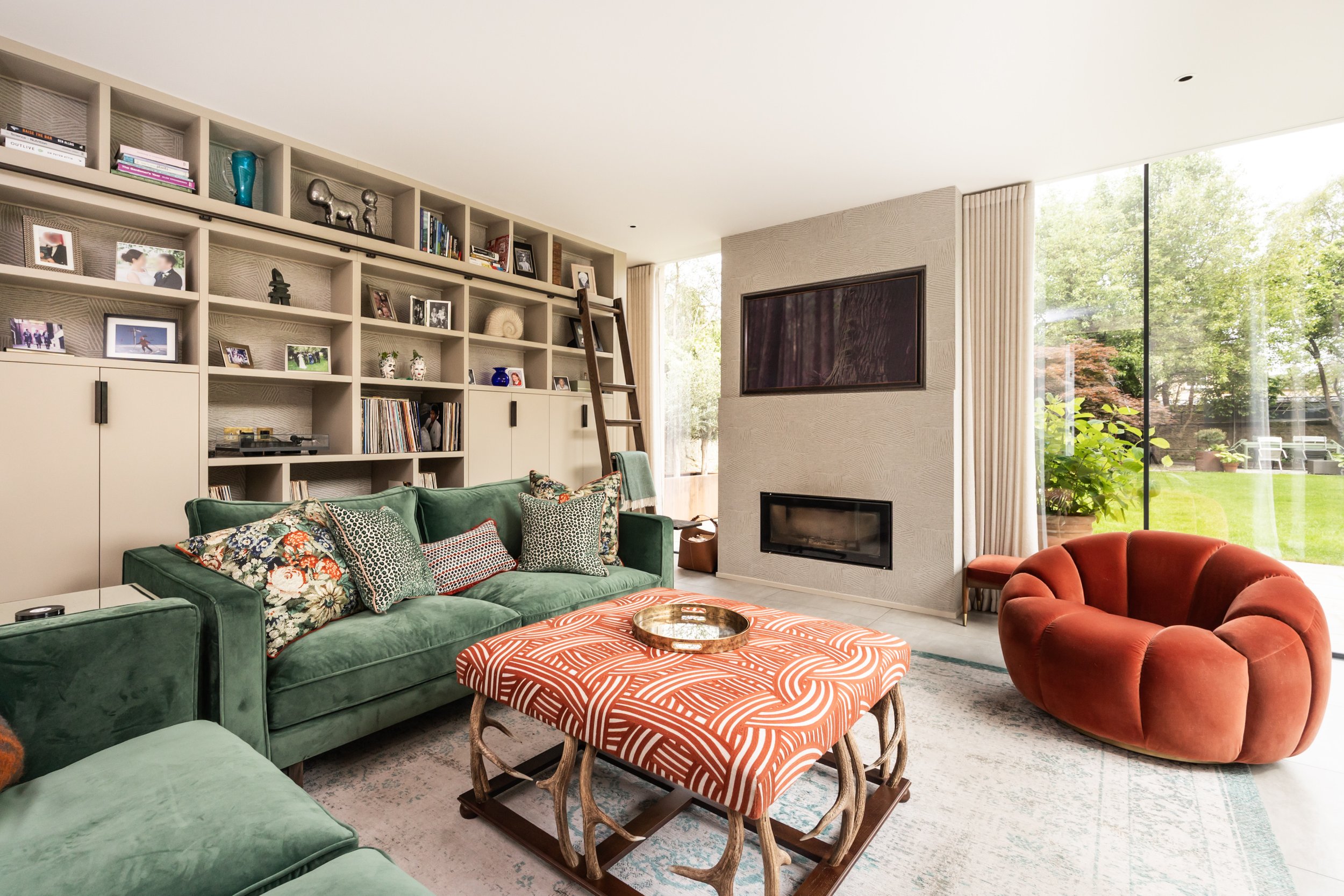 Living room with green sofa, patterned ottoman, red round chair, wood bookshelf with photos and books, white fireplace, large window showing garden, wall-mounted TV, ladder against bookshelf, beige curtains.