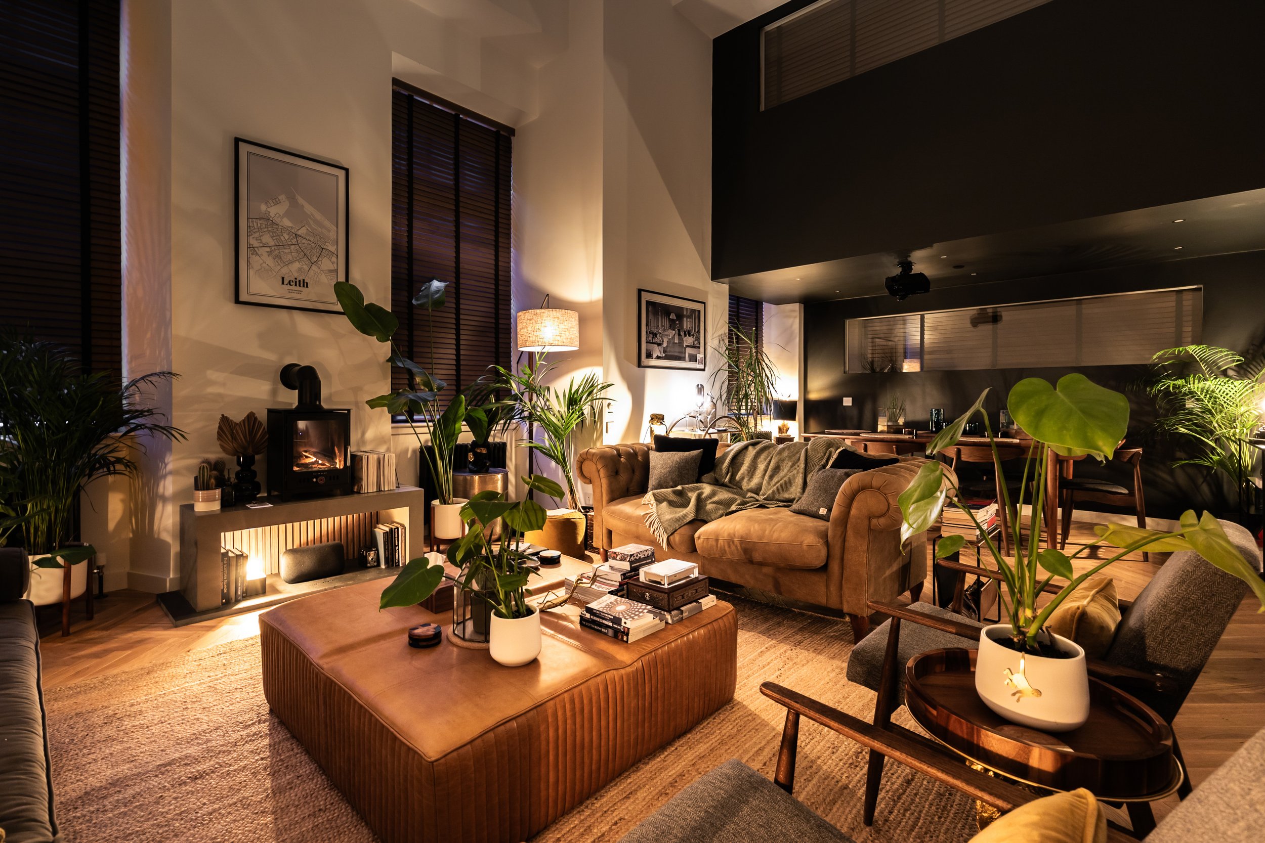 Living room with warm lighting, brown and gray furniture, plants, framed artwork, and a fireplace.