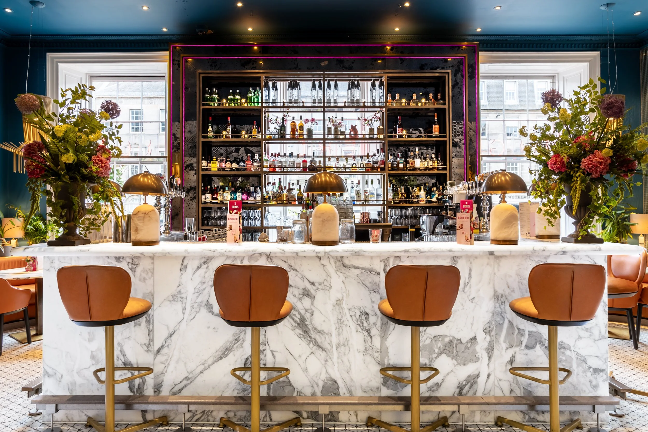 A modern bar with a white marble counter, four brown bar stools, and a backlit shelving unit filled with various bottles of alcohol. There are floral arrangements on either side of the bar, and large windows behind the shelves allowing natural light 