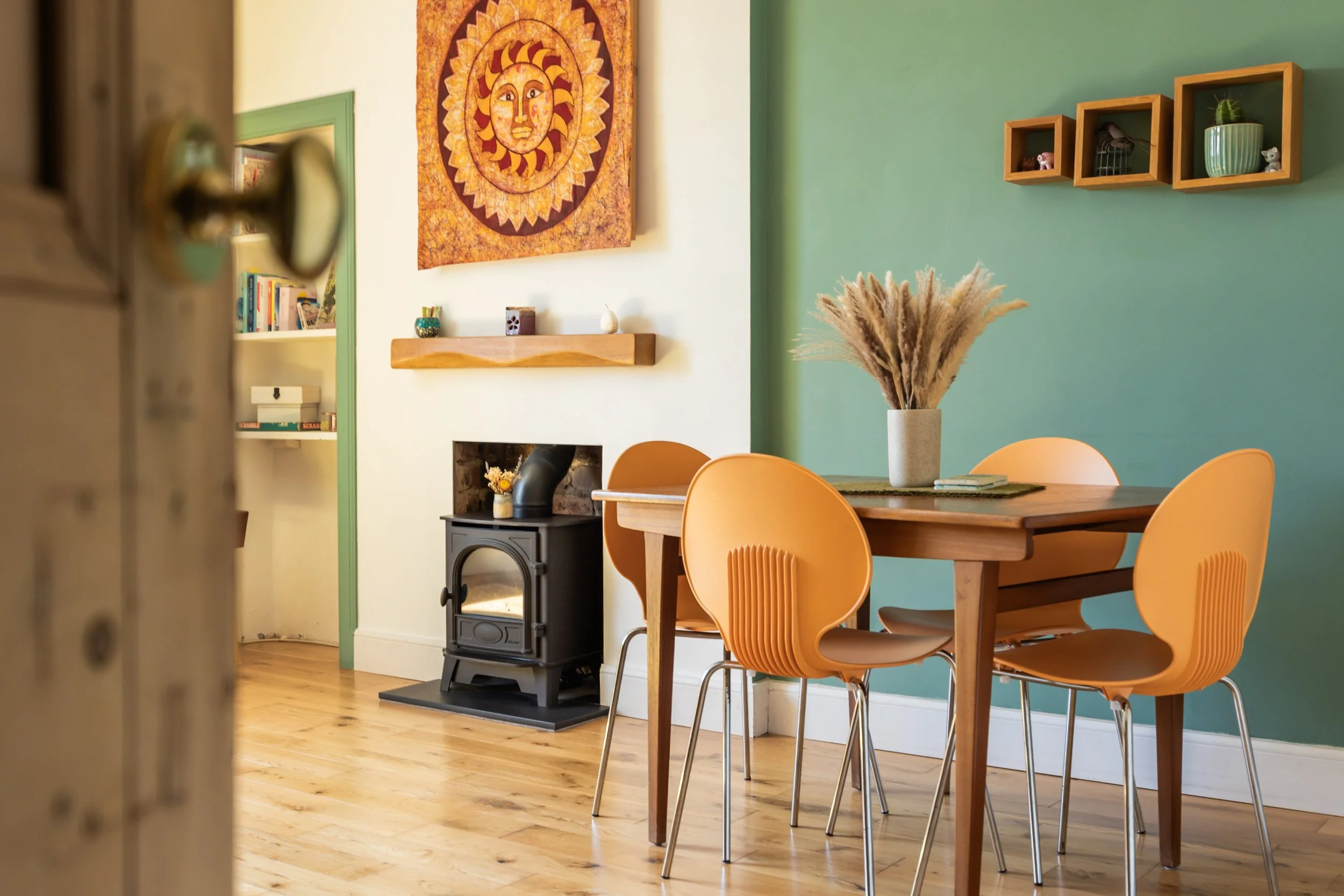 A cozy dining area with a wooden table and four orange chairs, a white vase with pampas grass on the table, a colorful sun and moon tapestry on the wall, a small wood stove, and decorative shelves with plants and small items.