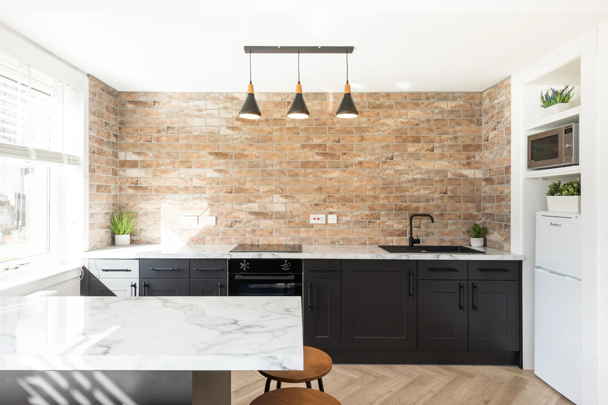 Modern kitchen with exposed brick wall, white marble countertops, black cabinets, black sink, and pendant lighting.