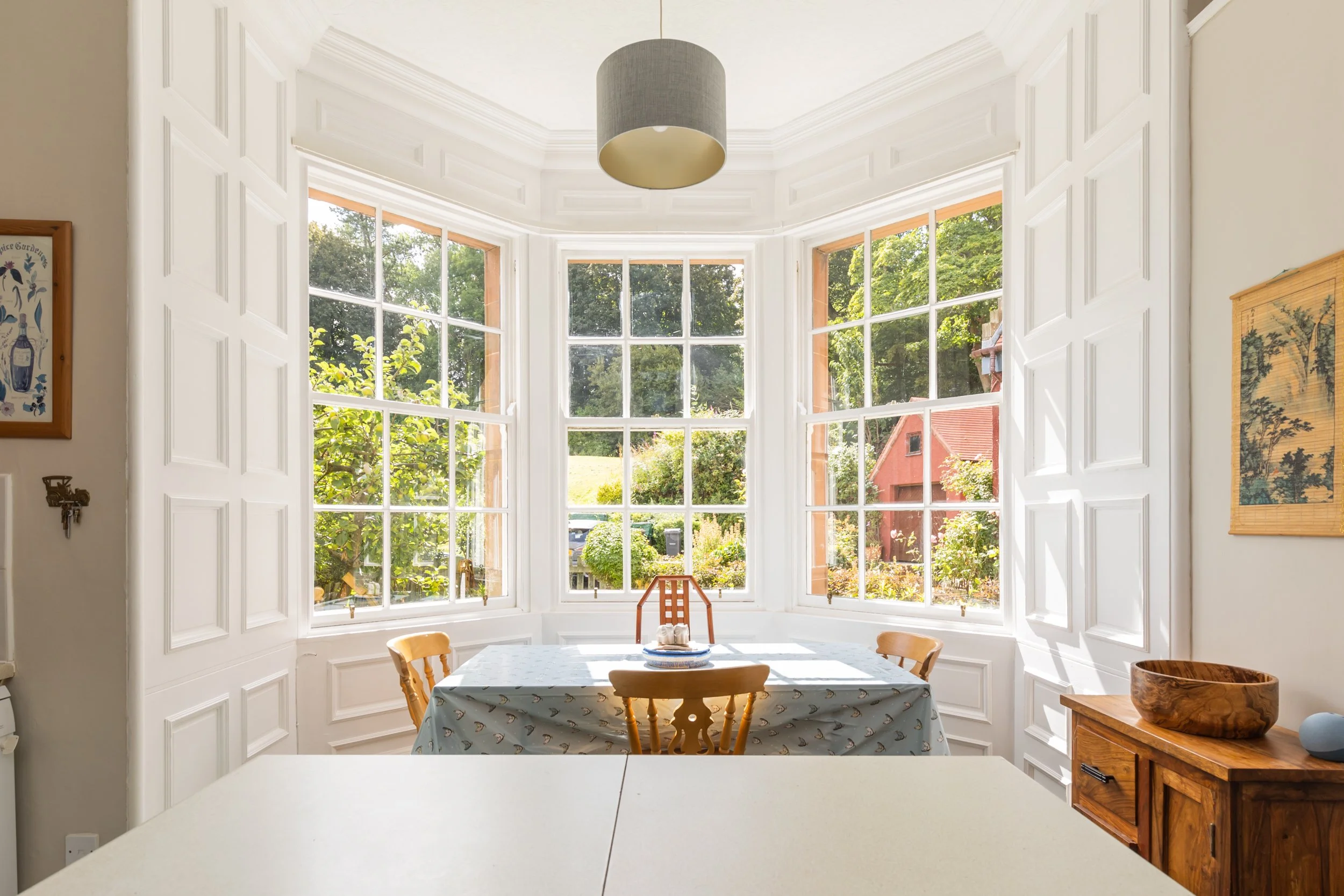 Sunlit dining area with a table covered by a patterned tablecloth, surrounded by wooden chairs, and large bay windows showing a lush green outdoor landscape.